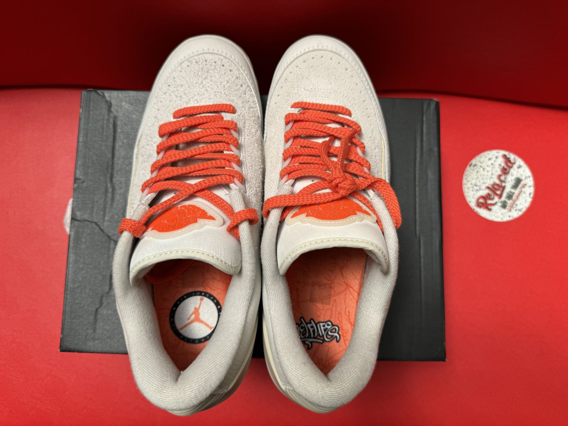 Pair of light gray sneakers with orange laces, one shoe upside down showing the sole. Shoes sit atop a black box on a red surface.