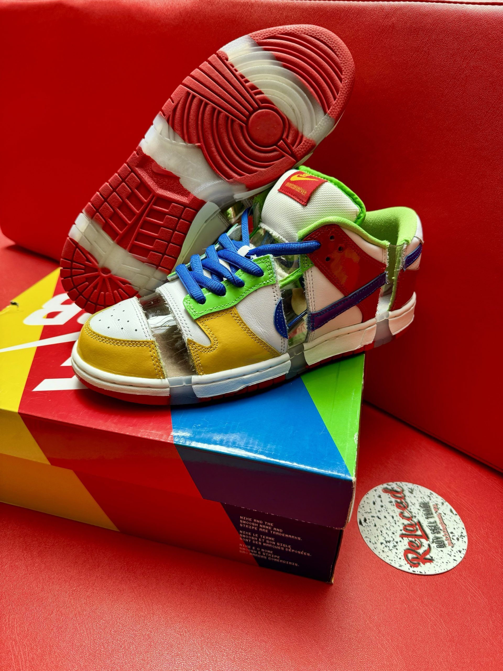 Nike Dunk Low SB sneakers, multi-colored patchwork design, sitting on box, red sole visible, red background.