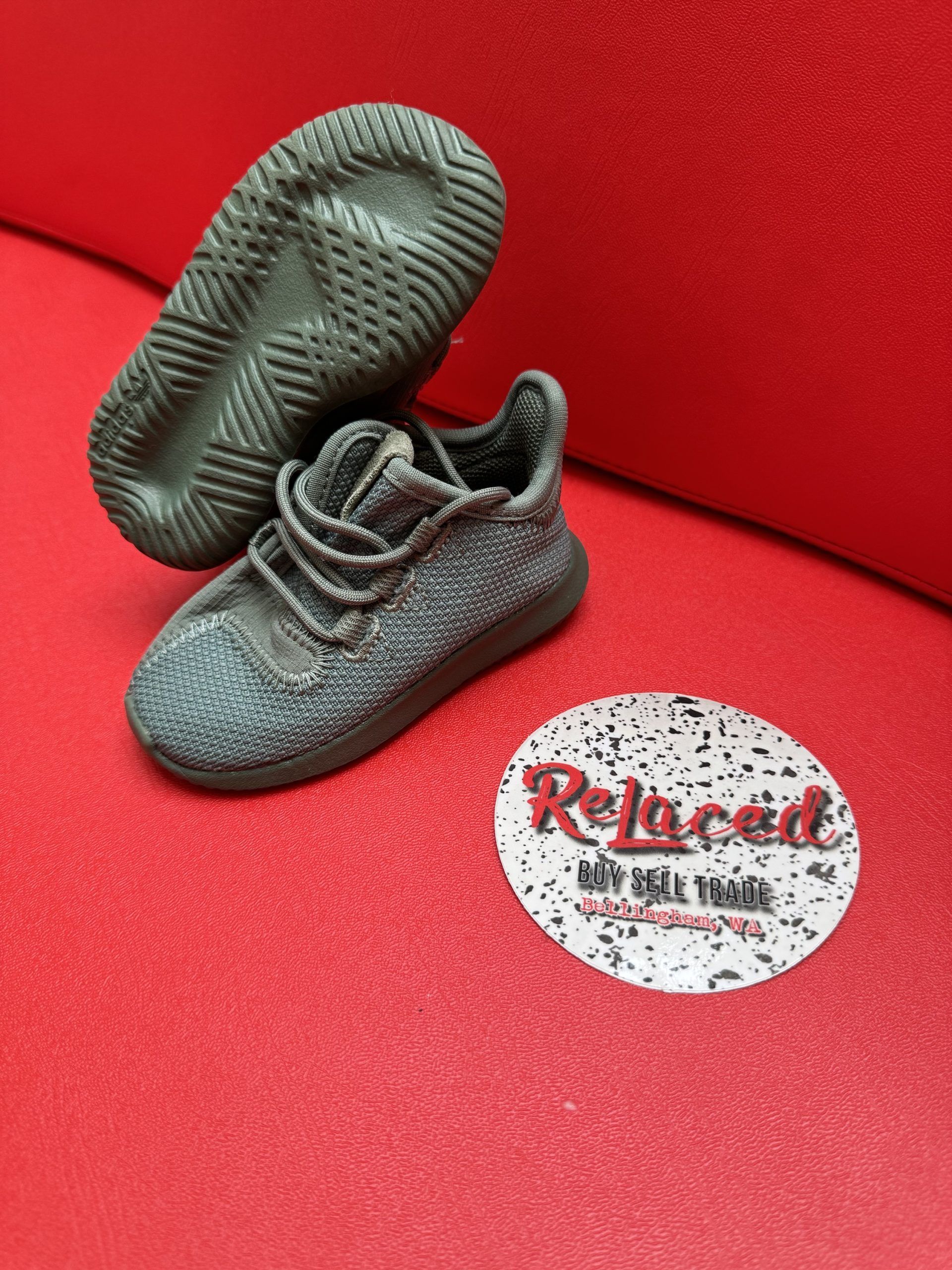 Small gray Adidas shoe on red surface next to a circular sticker that reads 