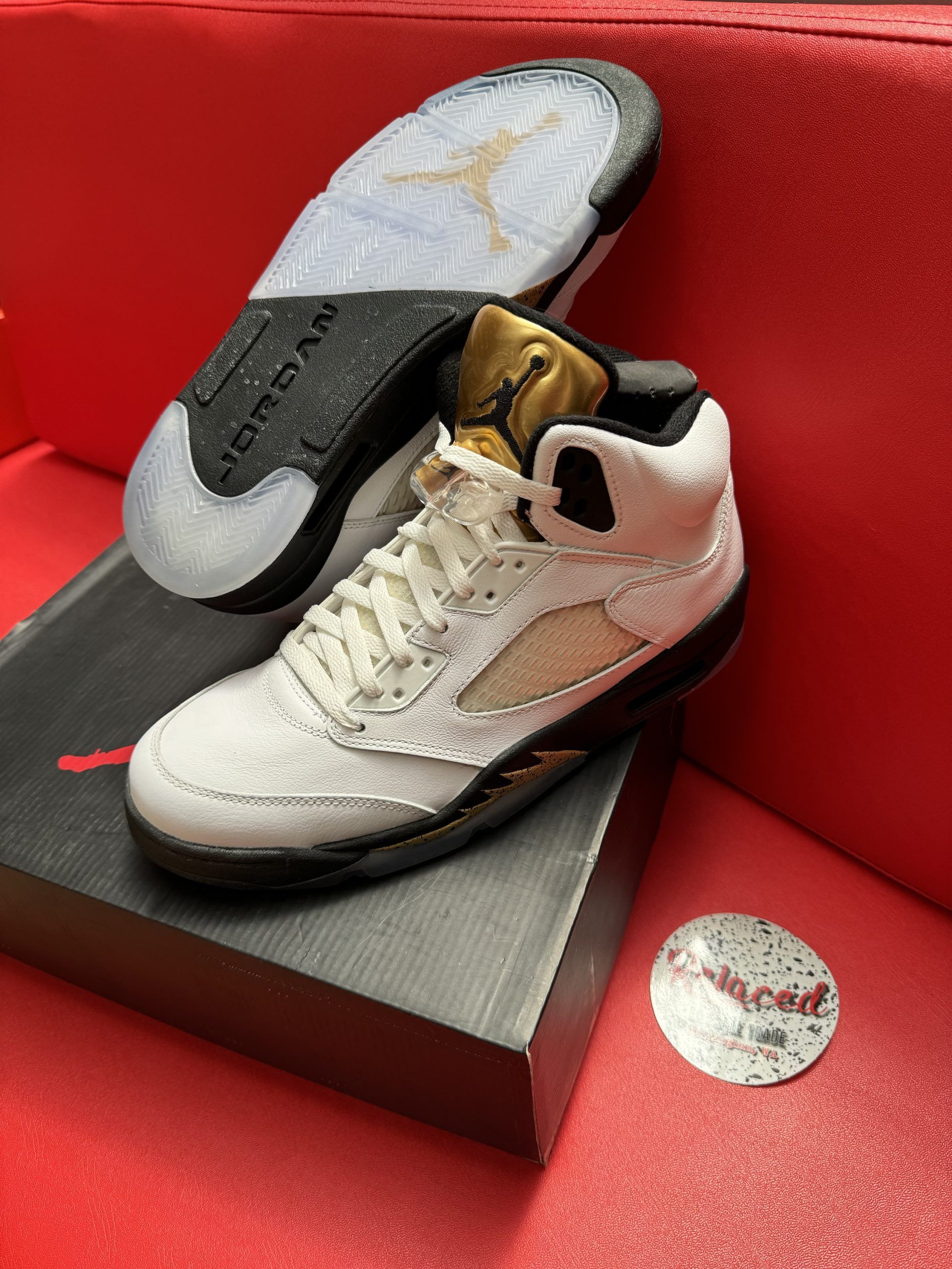 White and gold Air Jordan 5 sneakers with a black sole, on a red surface, near a shoe box.