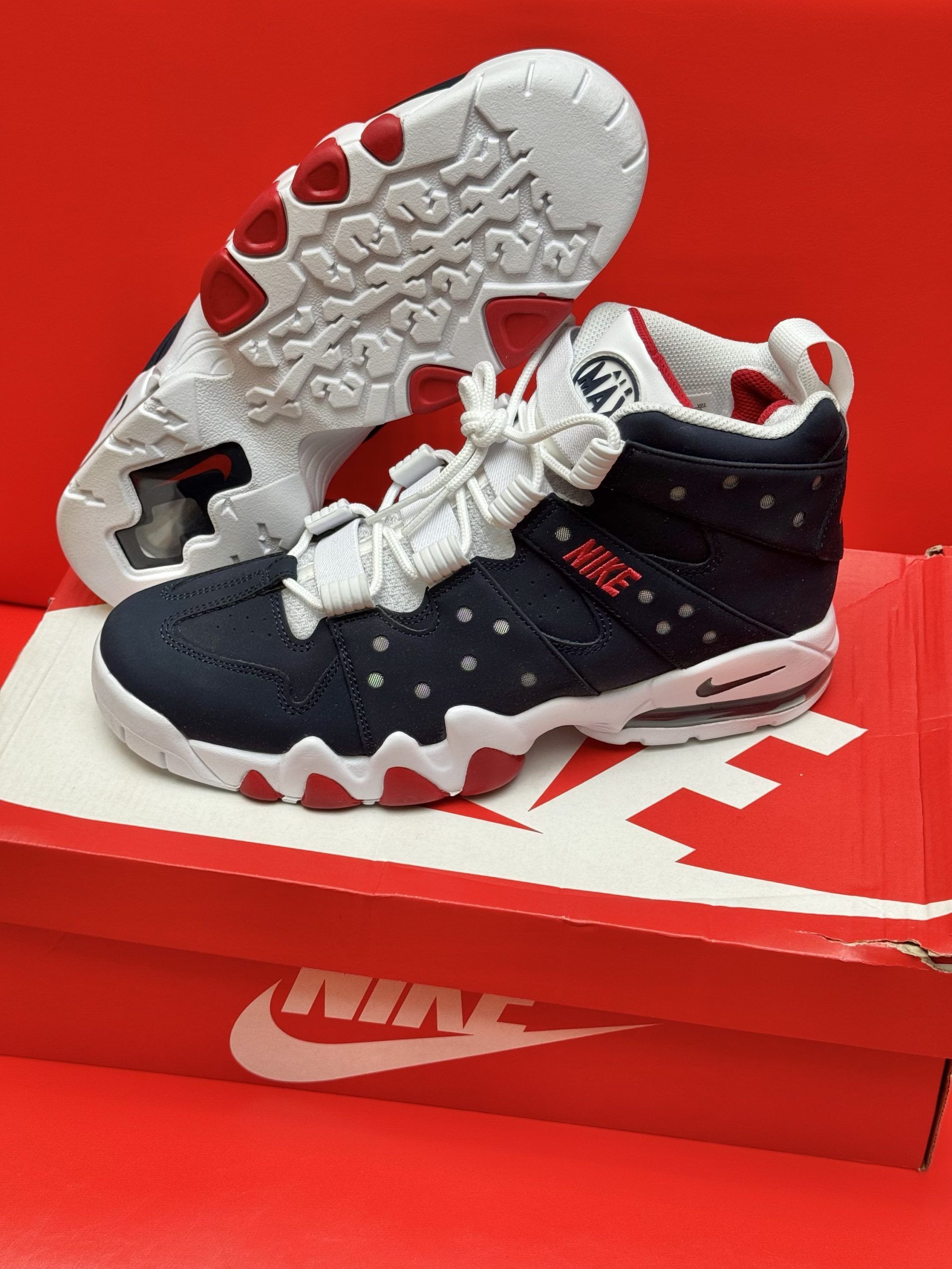 Navy and red Nike Air Max Barkley sneakers on a red Nike box, against a red backdrop.