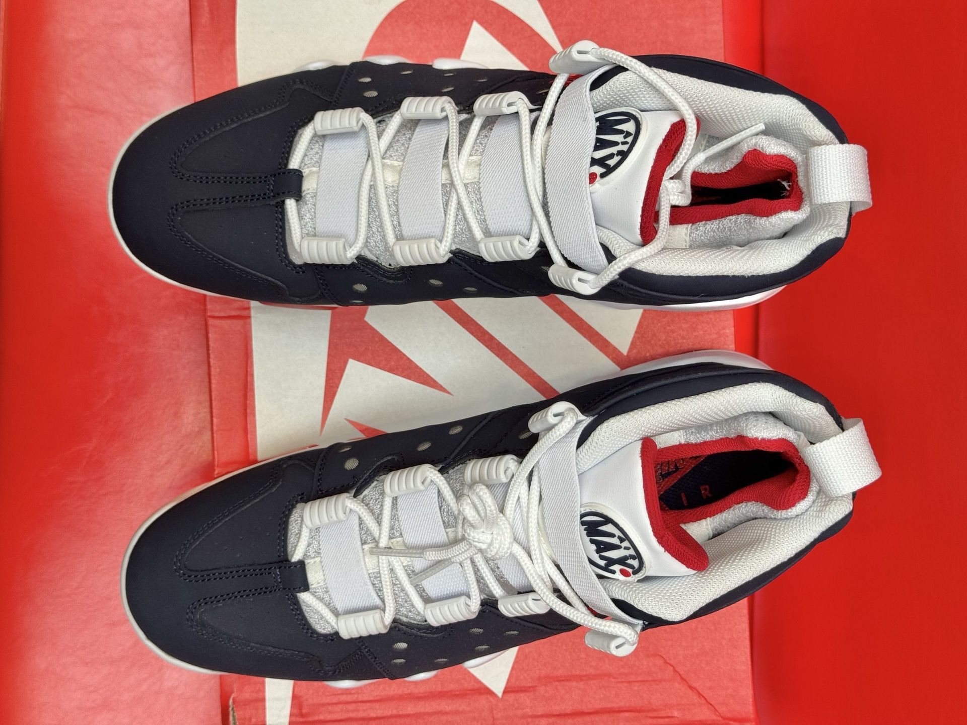 Navy and red Nike Air Max Barkley sneakers on a red Nike box, against a red backdrop.