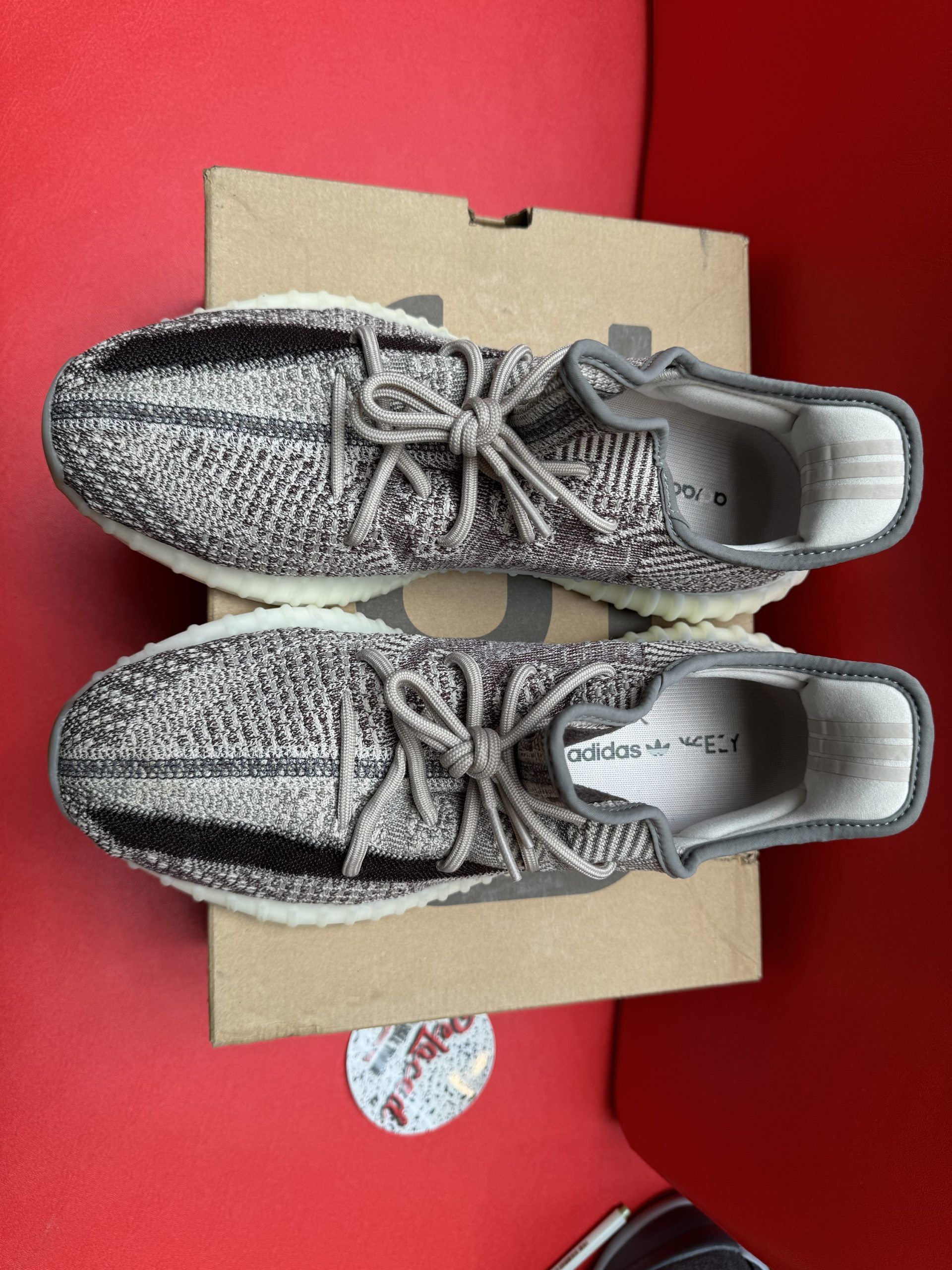 Pair of Adidas Yeezy Boost 350 V1 sneakers, brown and black pattern, on a box, red surface.