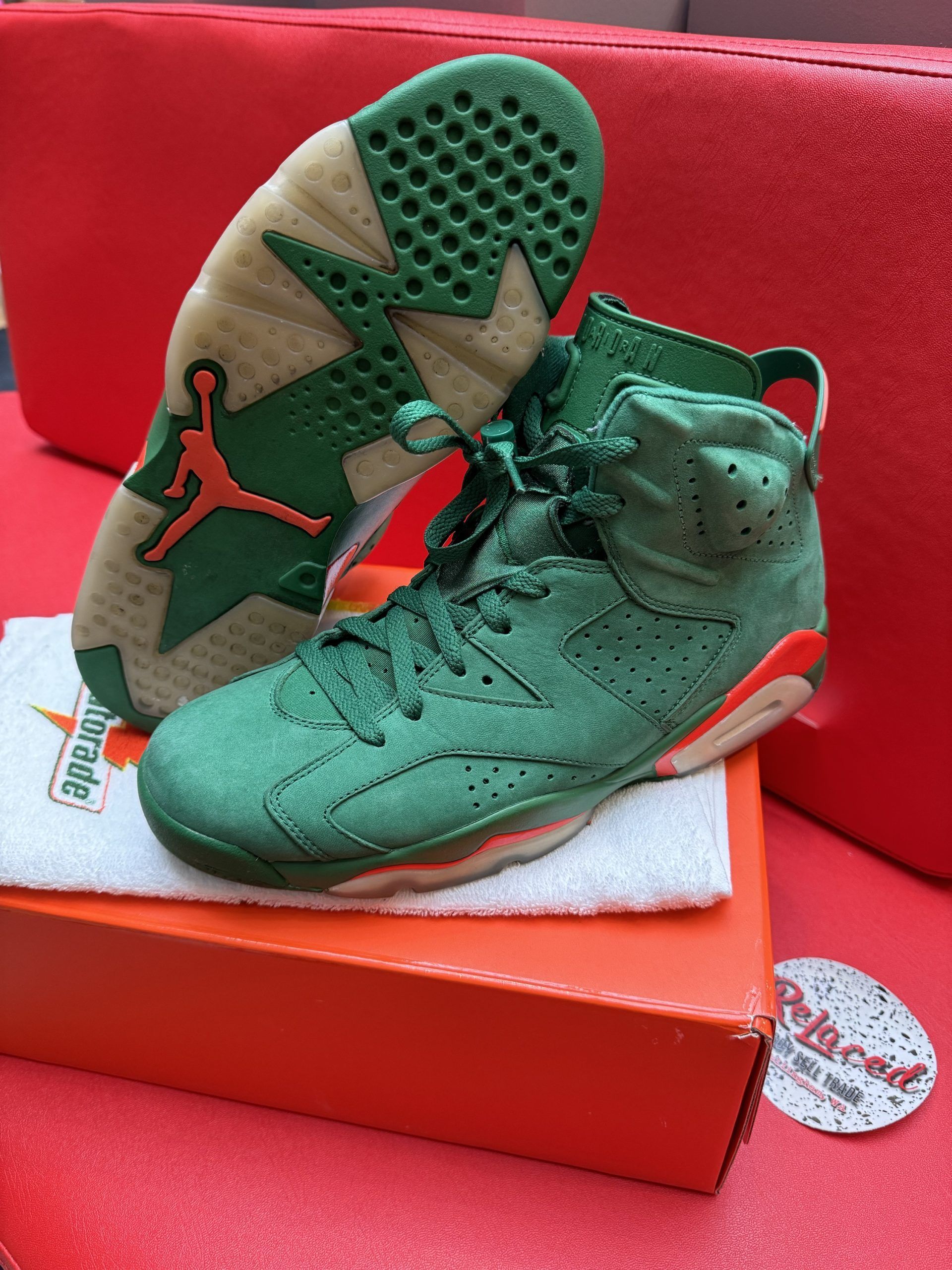 Green Air Jordan 6 sneakers, one upside down, resting on a box. Orange accents, red background.
