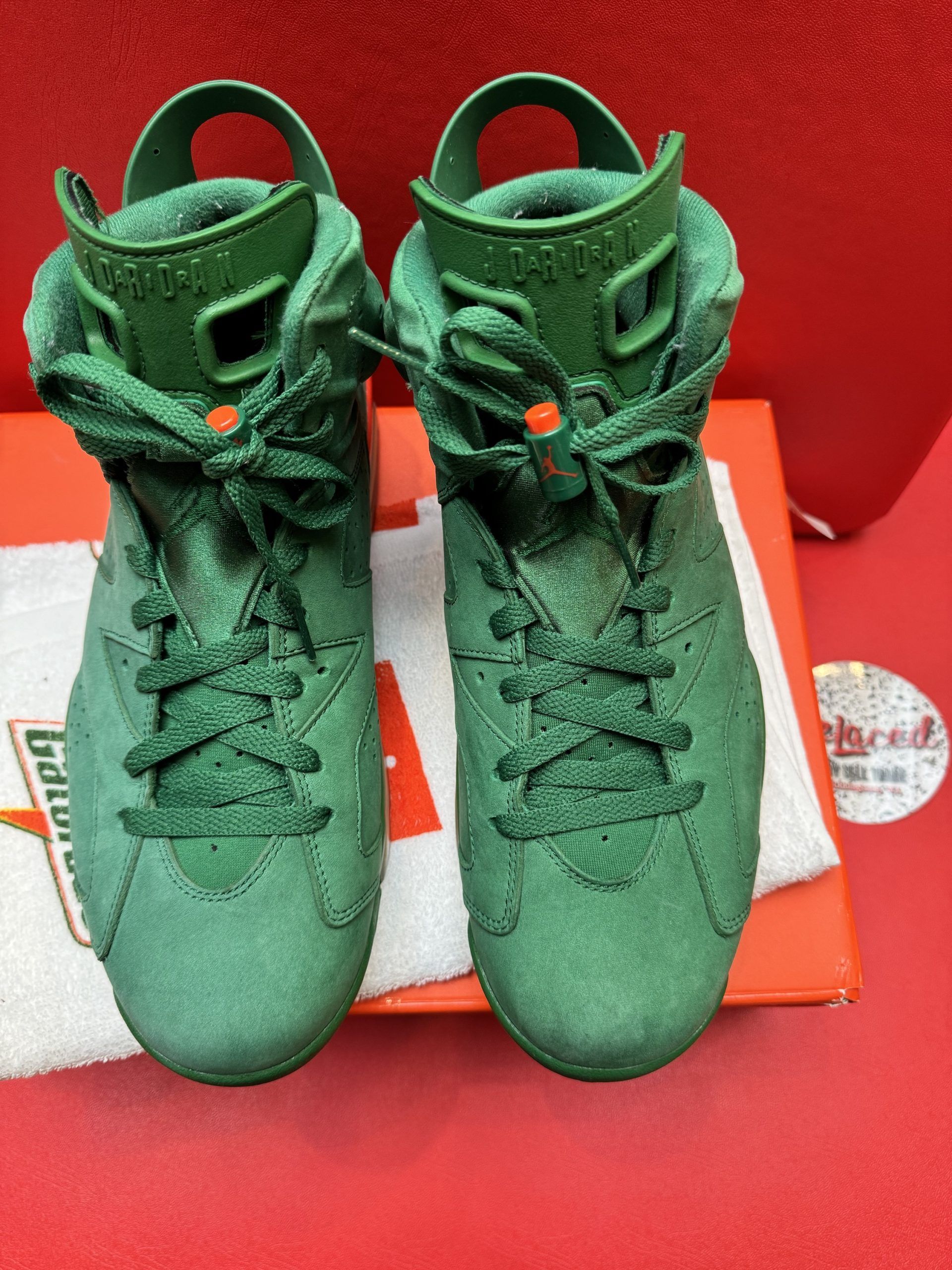 Green Air Jordan 6 sneakers, one upside down, resting on a box. Orange accents, red background.