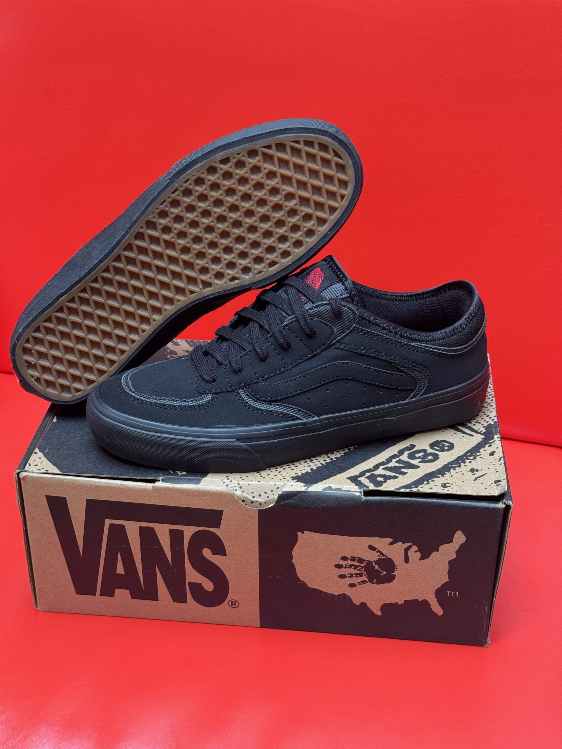 Black Vans sneakers on a shoebox with a brown sole, red background.