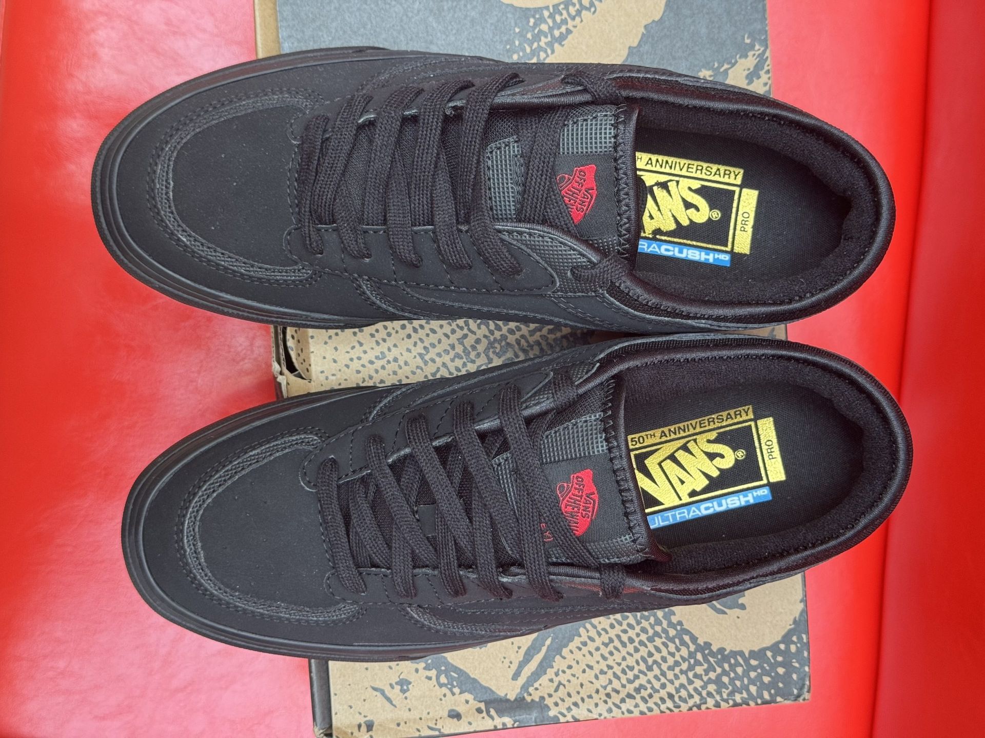 Black Vans sneakers on a shoebox with a brown sole, red background.