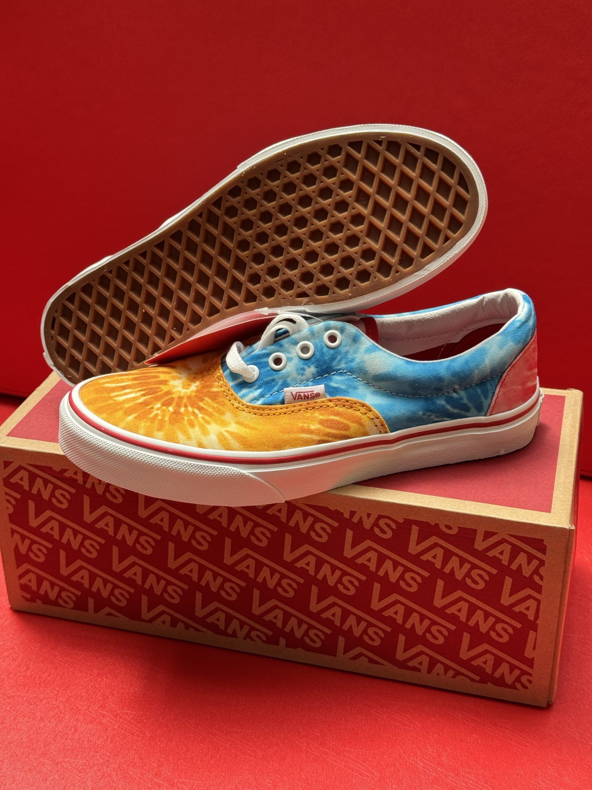 Vans sneakers in colorful tie-dye print on a box, with the sole visible. Red background.