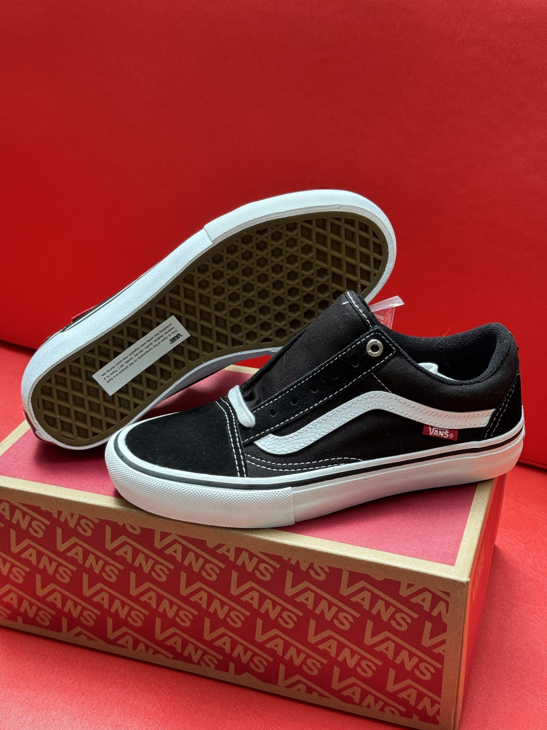 Black and white Vans sneakers on a box against a red backdrop. One shoe upside down showing the sole.
