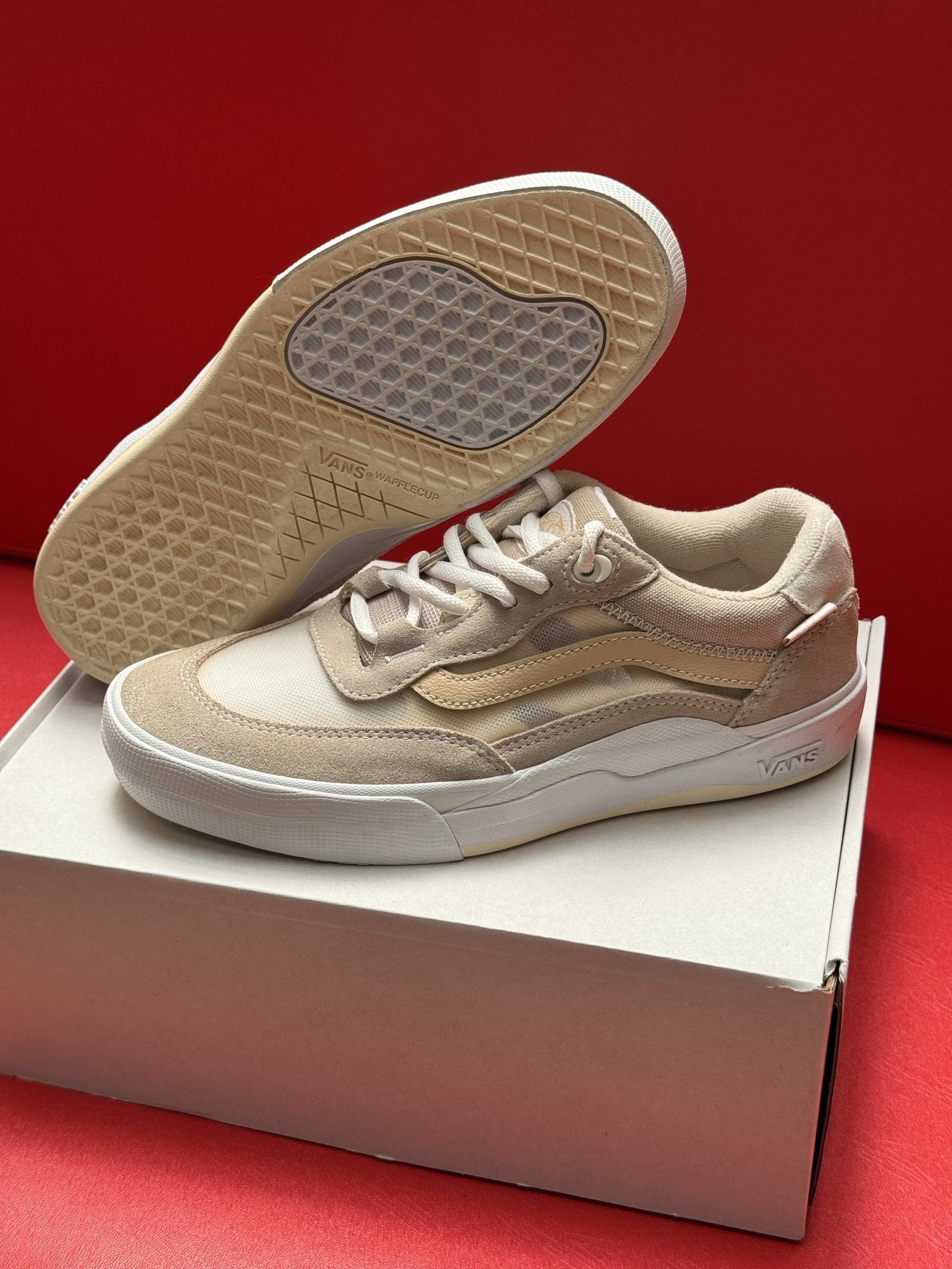 Tan and white Vans sneakers, one resting on a box, the other showing sole detail; red background.