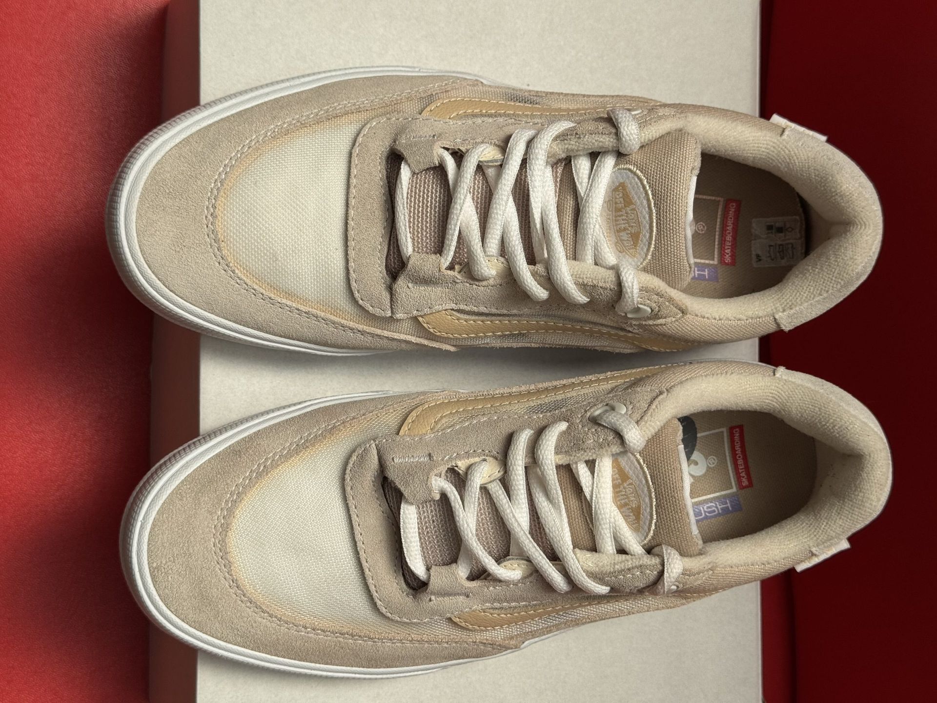 Tan and white Vans sneakers, one resting on a box, the other showing sole detail; red background.