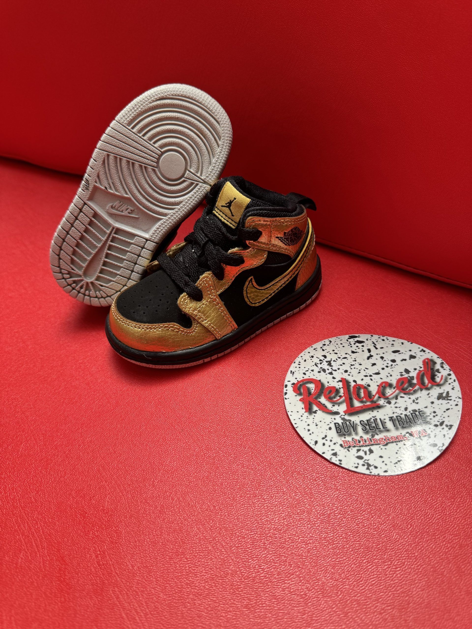 Gold and black baby Jordan sneaker with a red background, white sole is visible.
