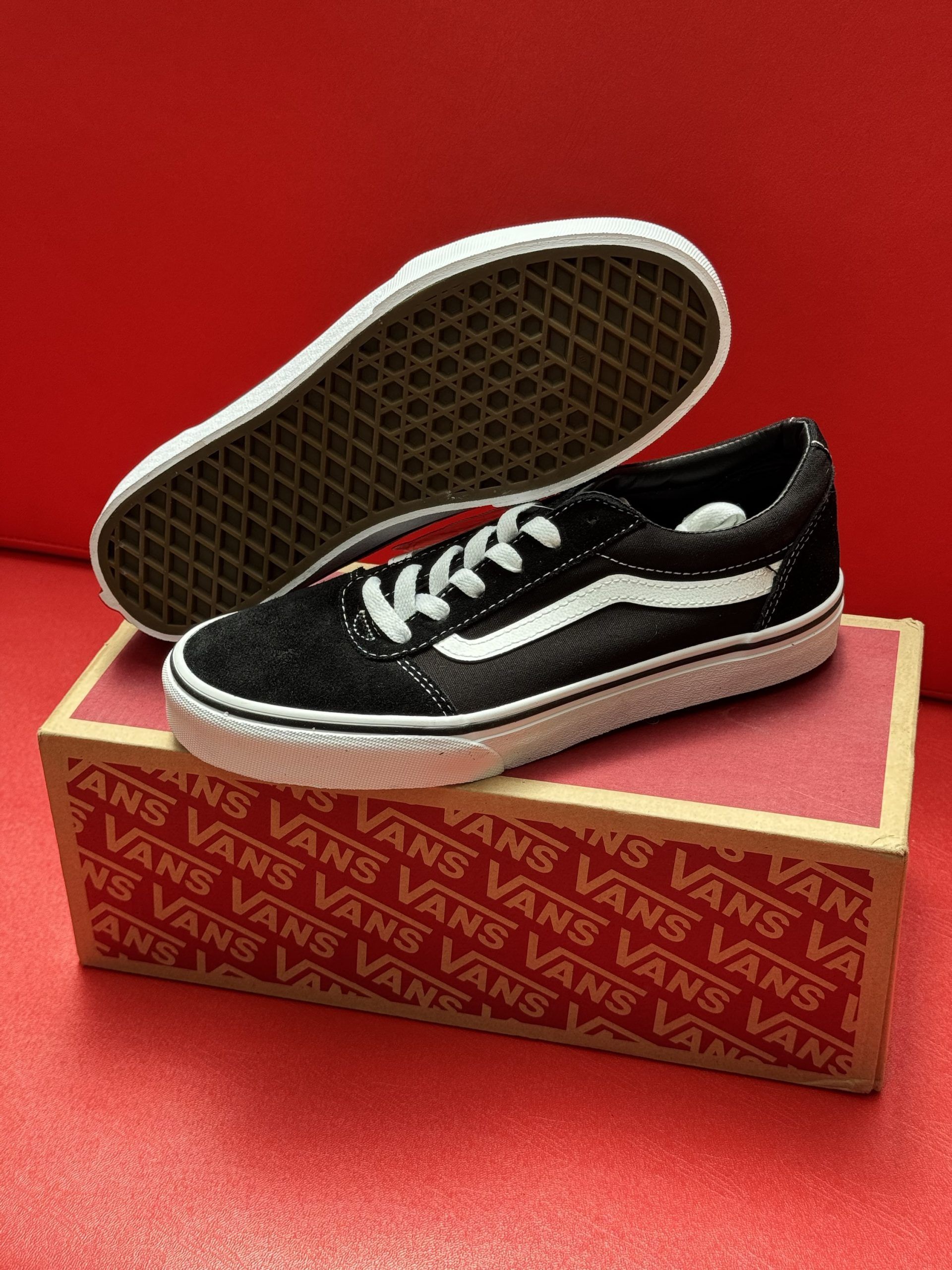 Black Vans sneakers with white accents and a box, on a red surface. The sole is visible.