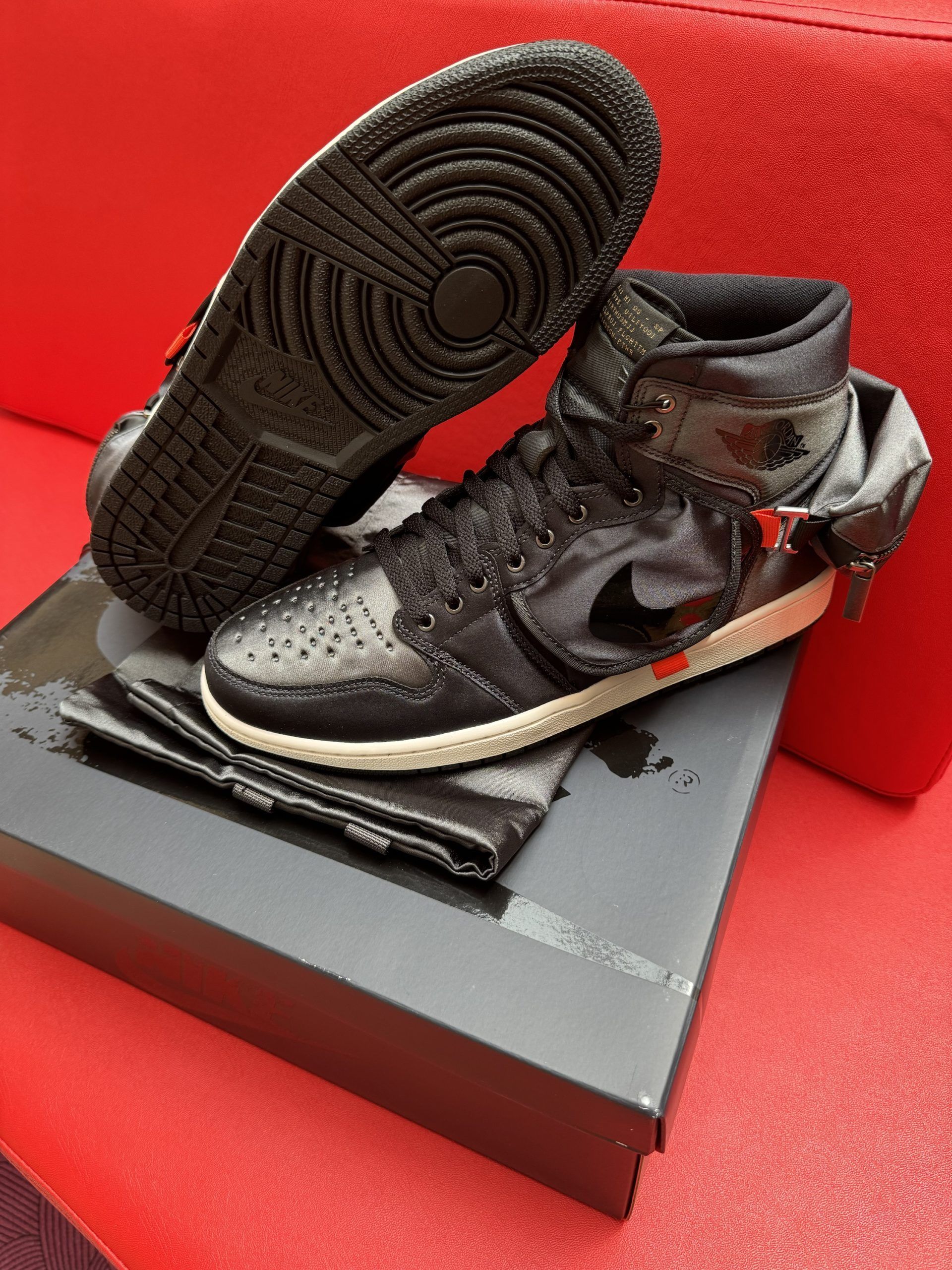 Black Air Jordan 1 sneaker on a box, with the sole facing up, on a red surface.