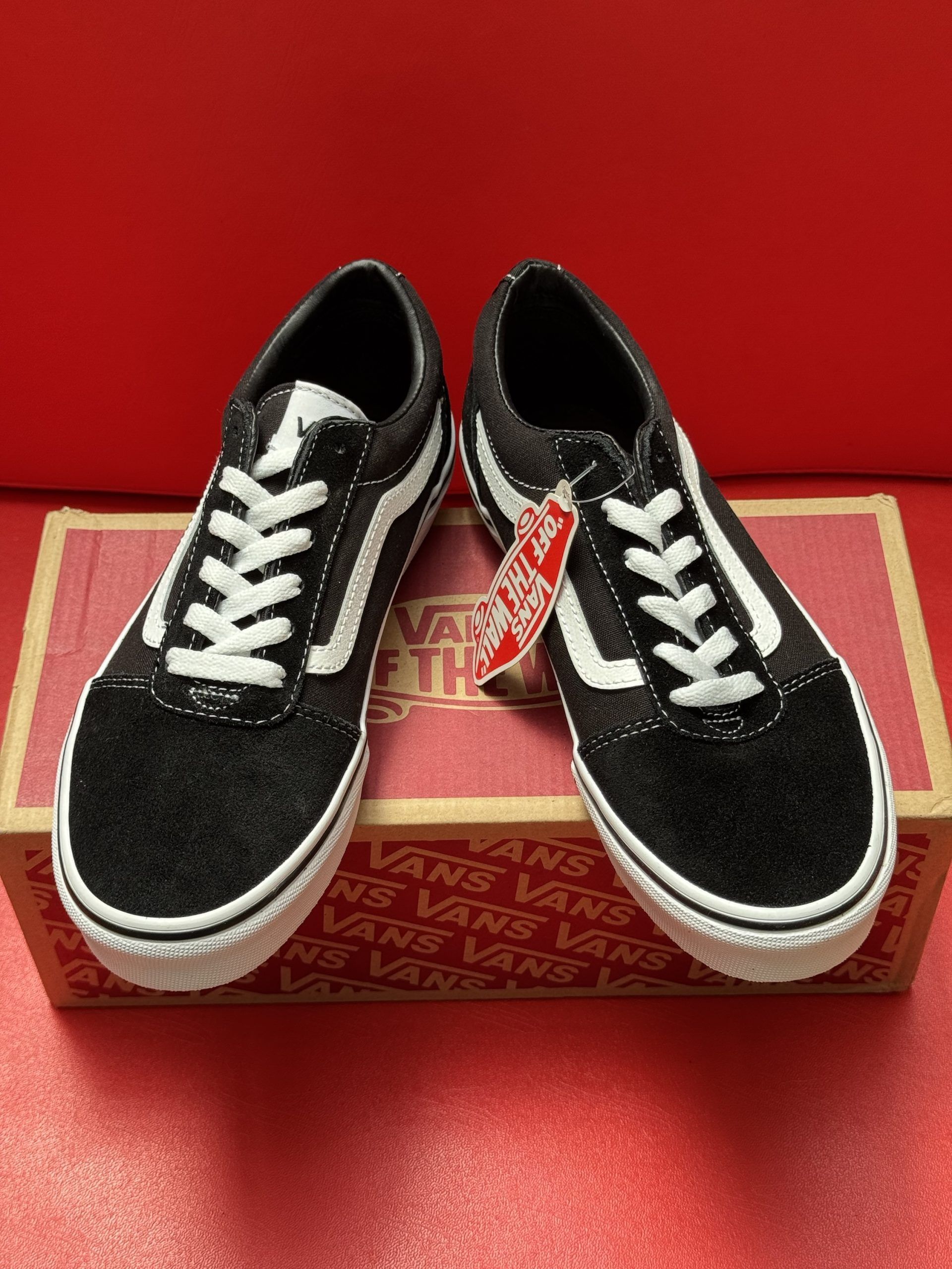Black Vans sneakers with white accents and a box, on a red surface. The sole is visible.