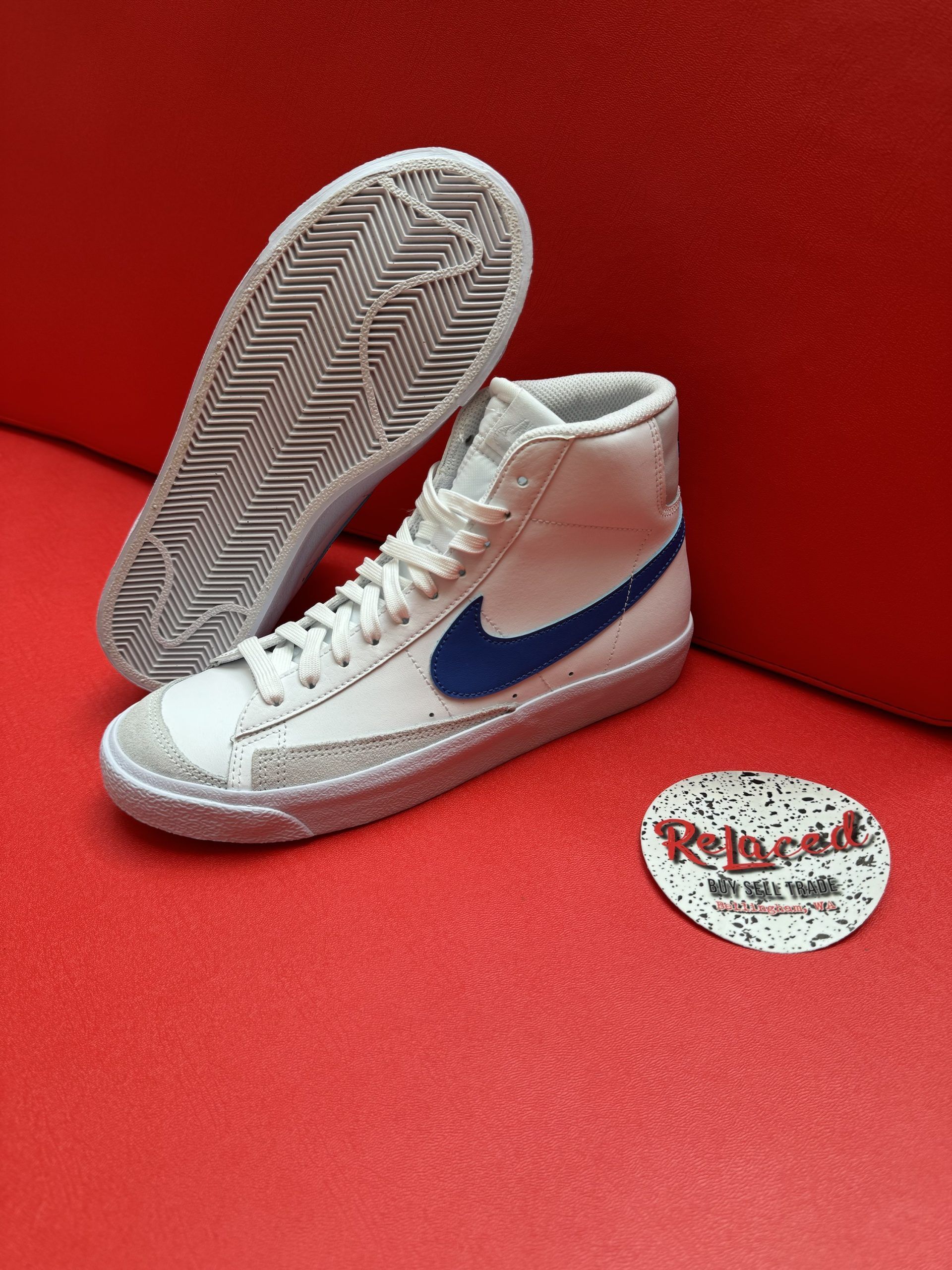 White Nike Blazer high-top sneaker with blue swoosh, resting on a red surface, with a sole showing.