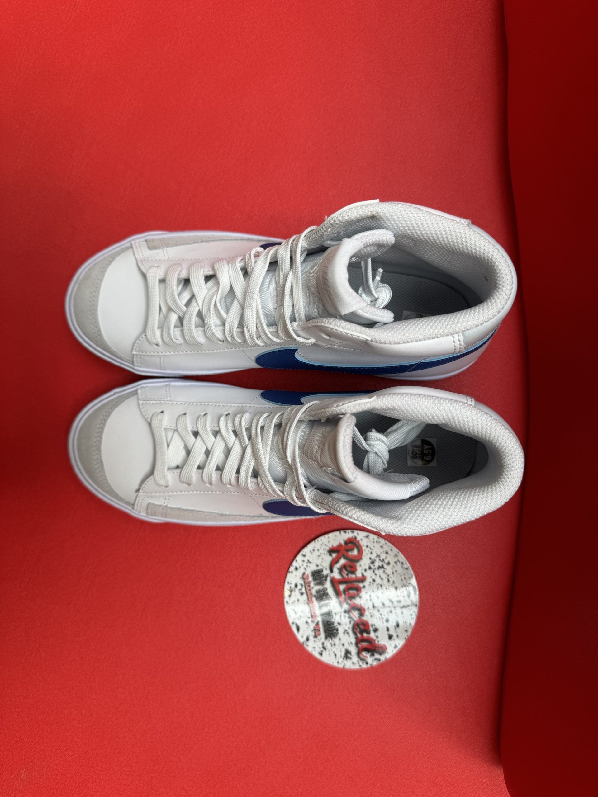 White Nike Blazer high-top sneaker with blue swoosh, resting on a red surface, with a sole showing.