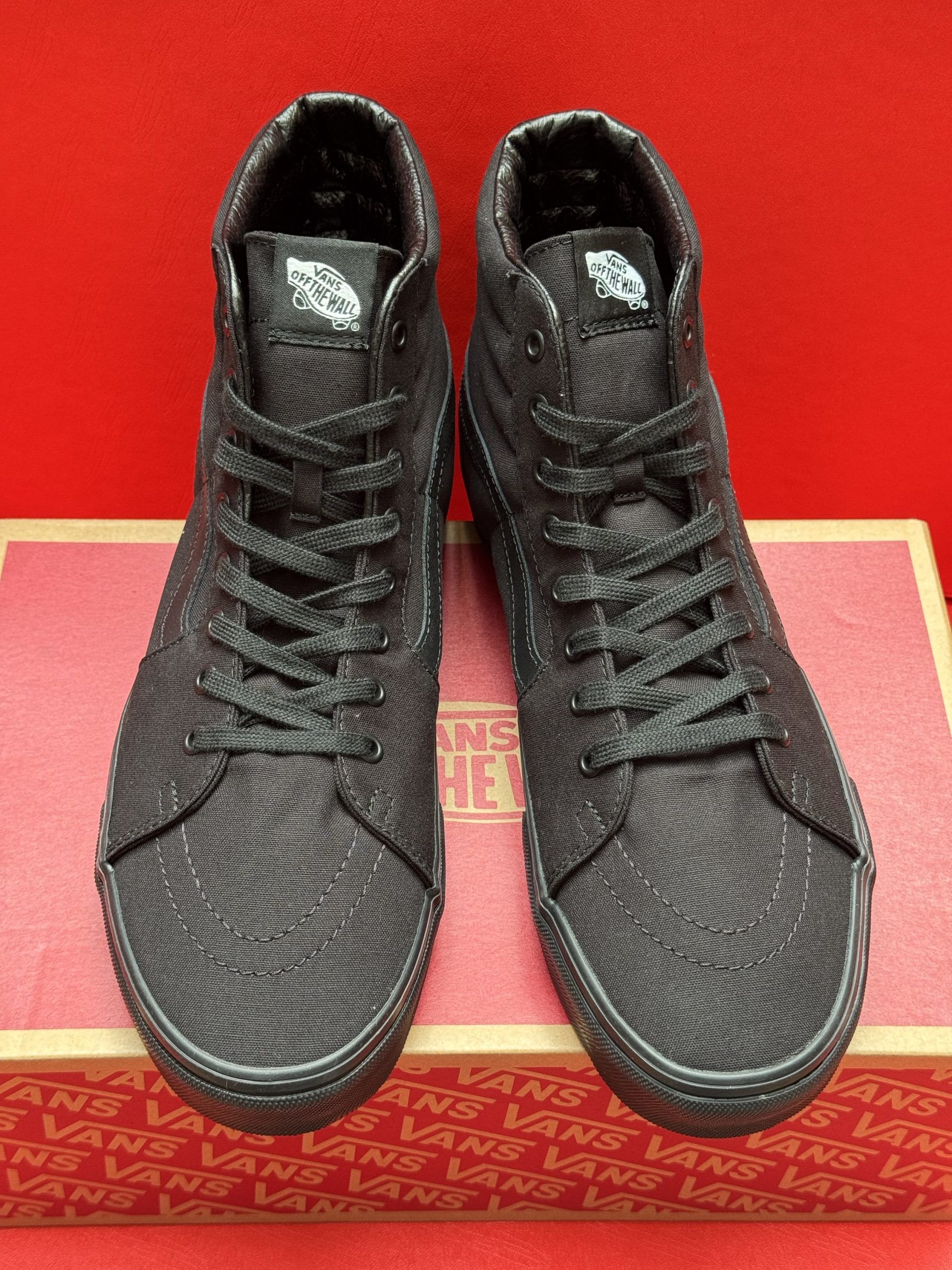 Black Vans Sk8-Hi sneakers on a red surface, with one shoe's sole visible. They are sitting on a Vans box.