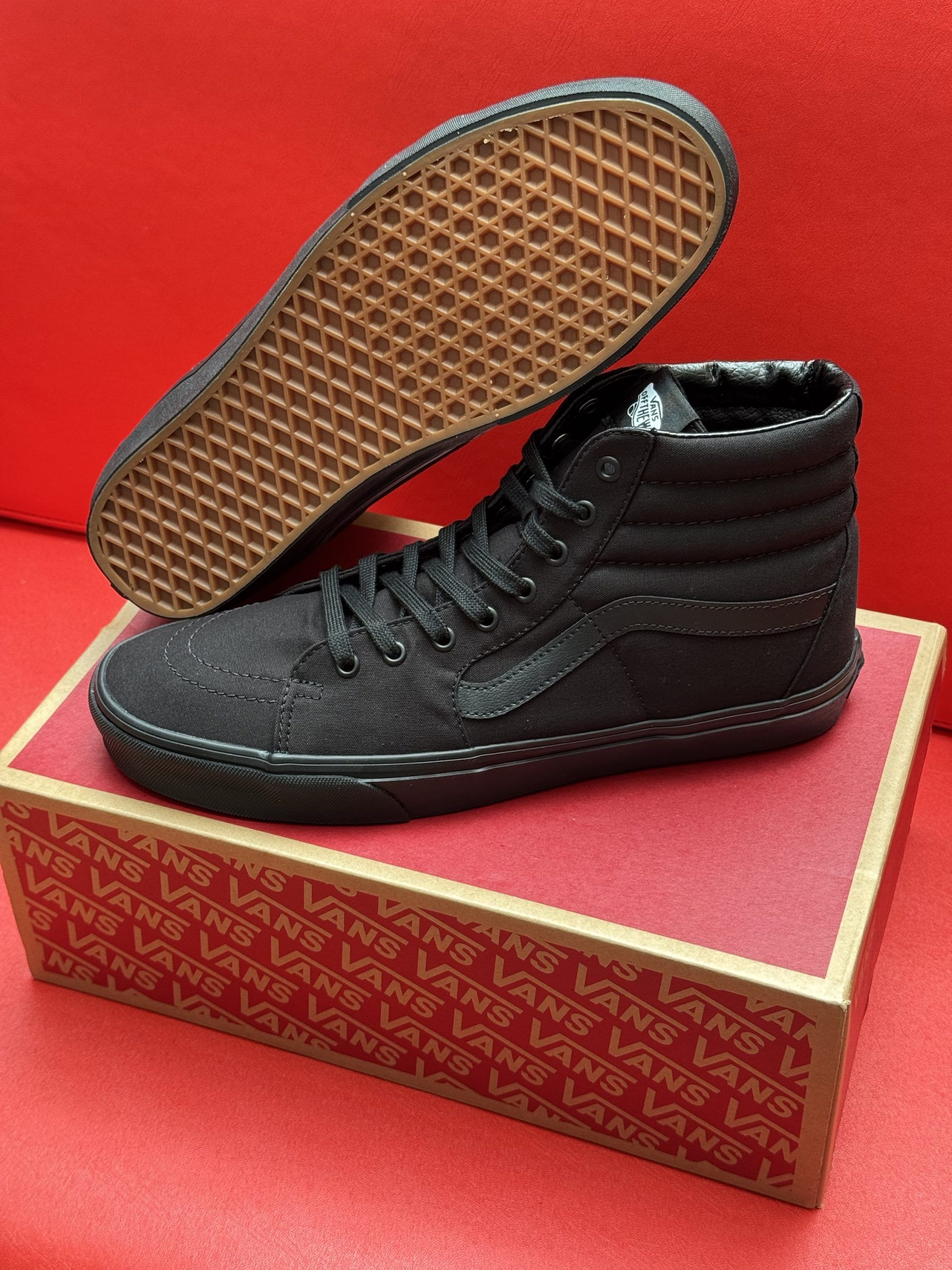 Black Vans Sk8-Hi sneakers on a red surface, with one shoe's sole visible. They are sitting on a Vans box.