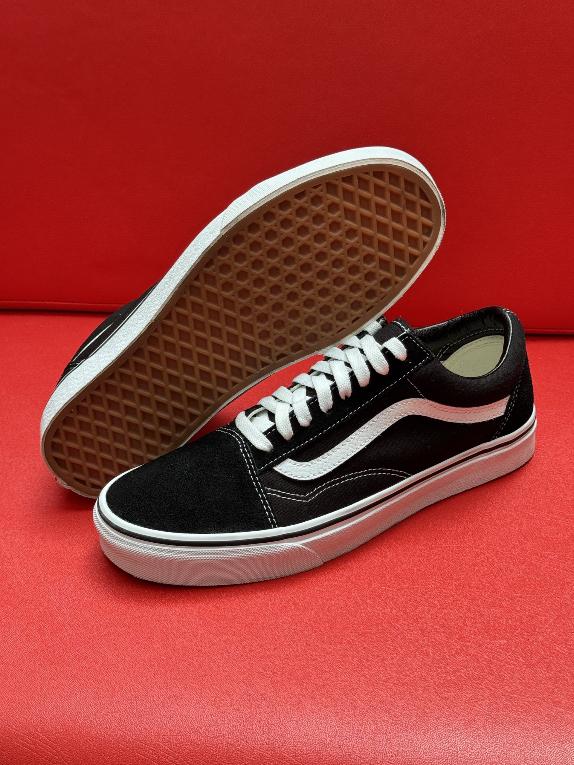 Black Vans Old Skool sneakers with white stripe and laces, on a red surface. One shoe is upside down, revealing the sole.