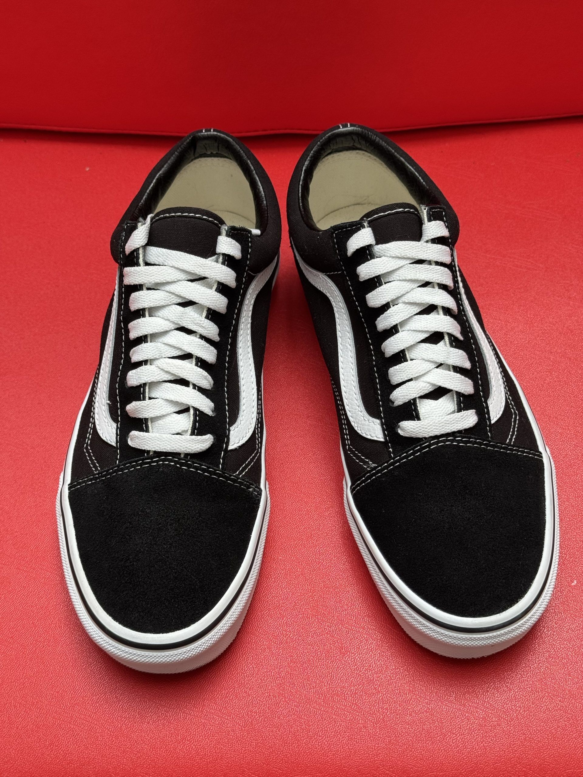 Black Vans Old Skool sneakers with white stripe and laces, on a red surface. One shoe is upside down, revealing the sole.