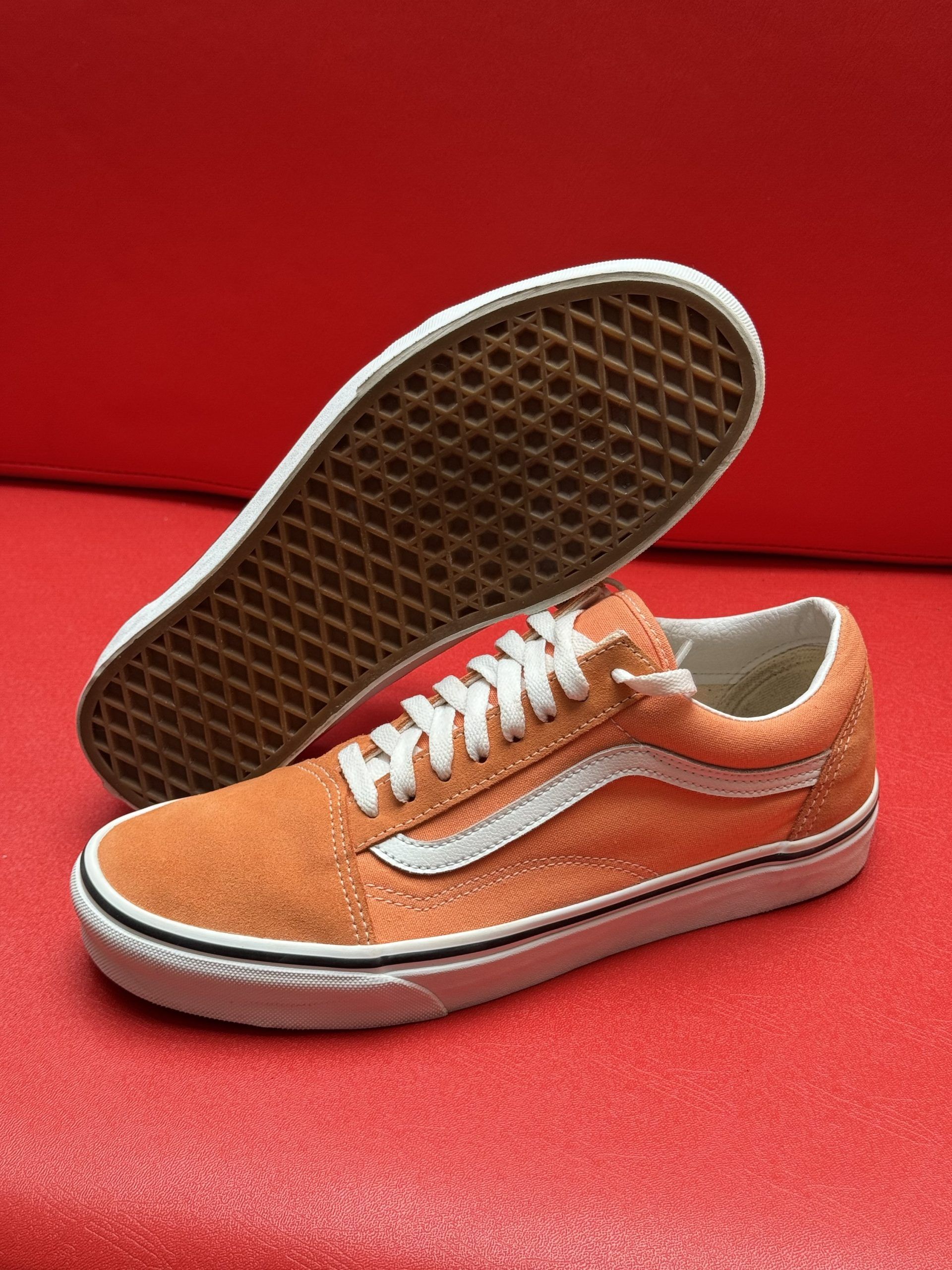 Orange Vans Old Skool sneakers with white stripe and laces on a red surface. Sole of one shoe visible.