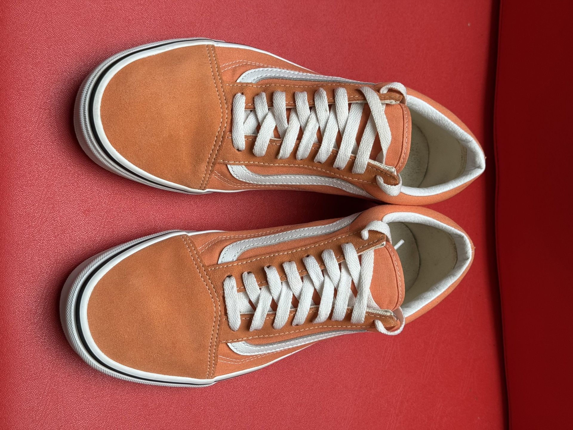 Orange Vans Old Skool sneakers with white stripe and laces on a red surface. Sole of one shoe visible.