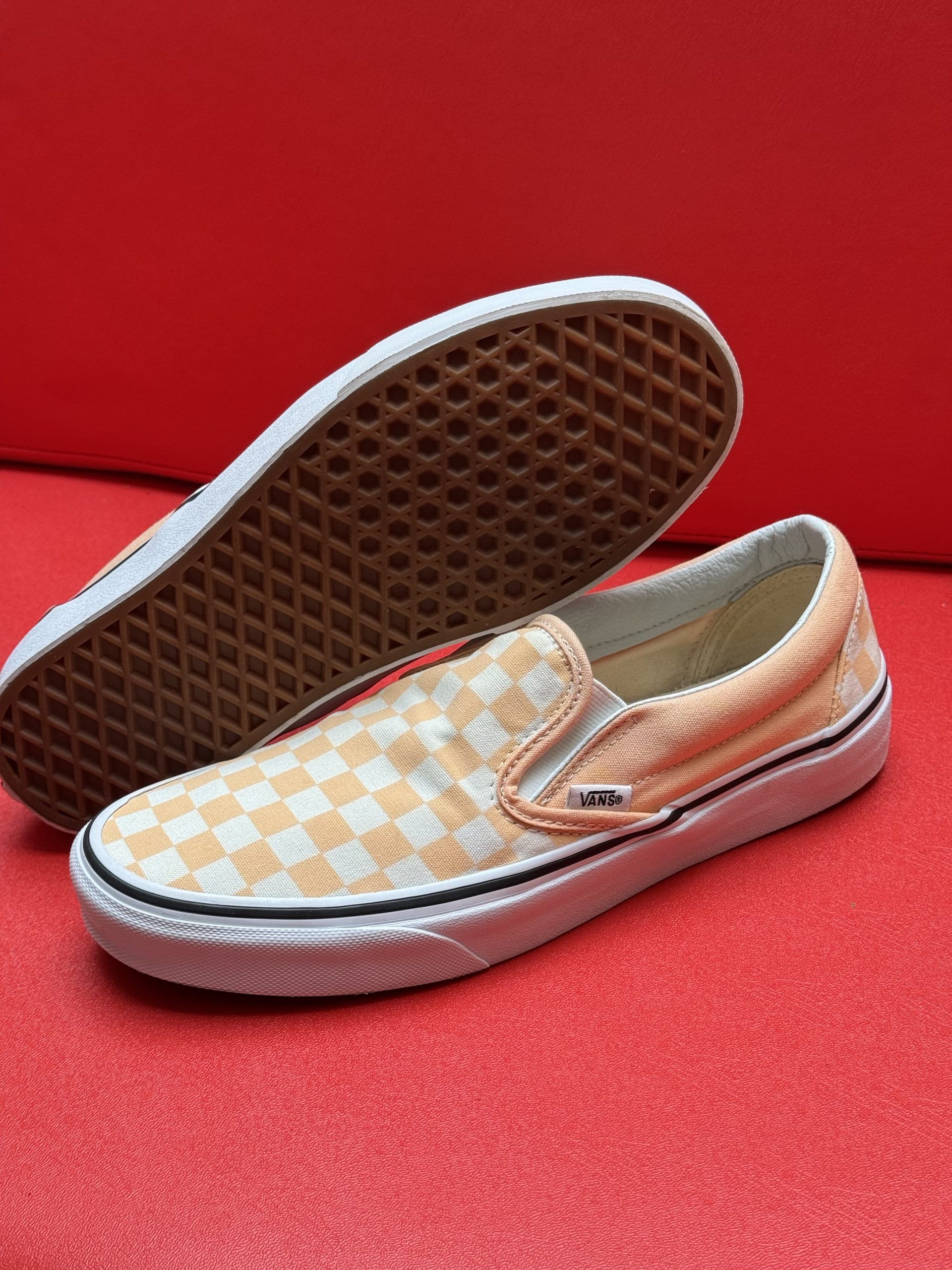 Vans slip-on shoes with orange and white checkered pattern, white soles, and a waffle-patterned brown sole, on a red surface.