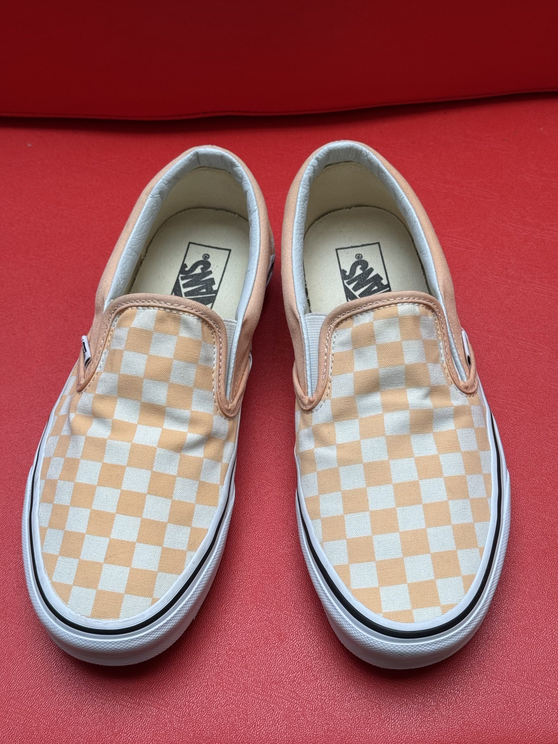 Vans slip-on shoes with orange and white checkered pattern, white soles, and a waffle-patterned brown sole, on a red surface.