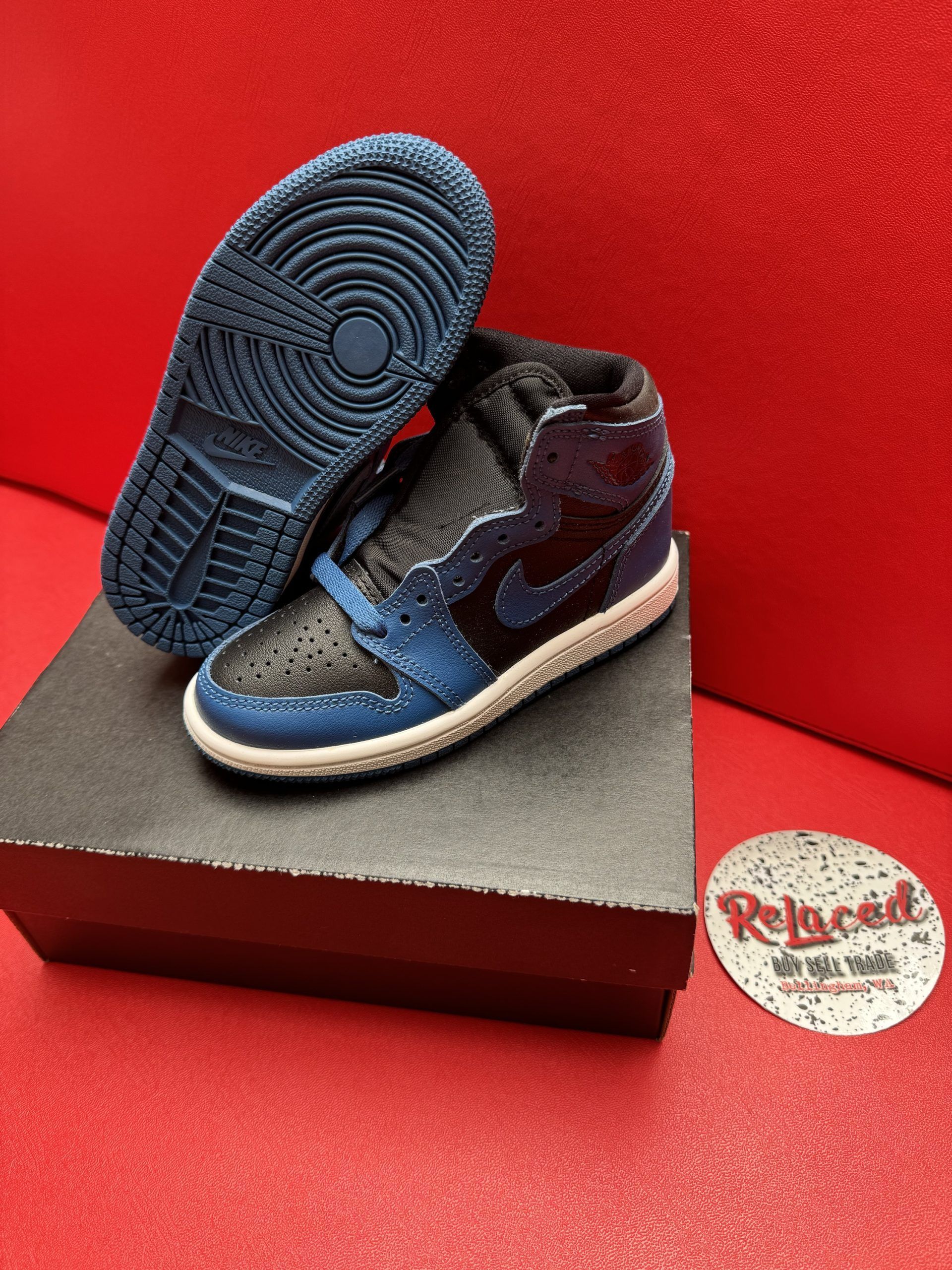 Blue and black Air Jordan 1 toddler shoes on a box, sole up. Red background.