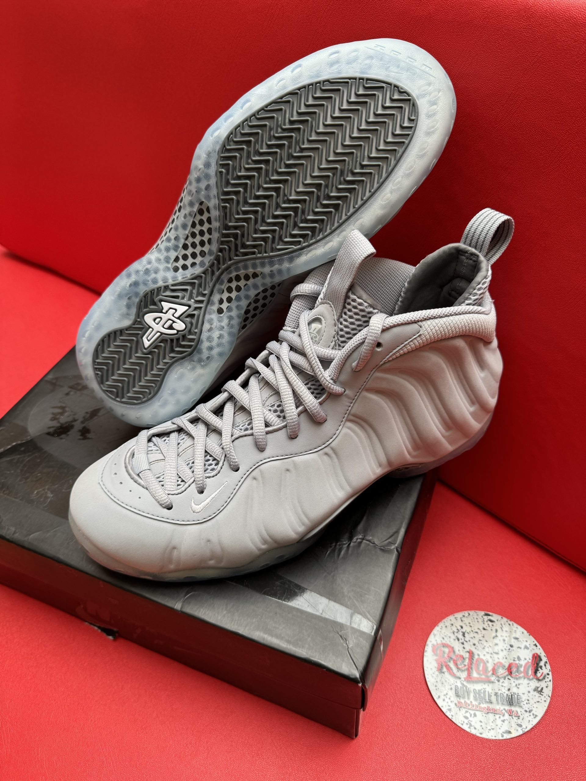 Gray Nike Foamposite sneakers on a box, with a view of the sole, resting on a red surface.