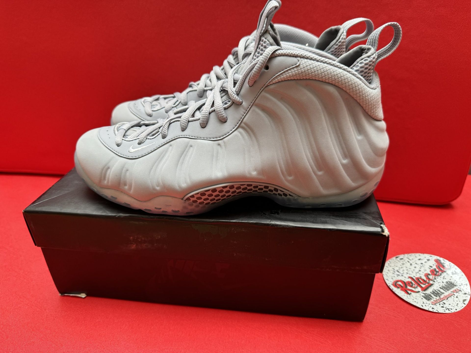 Gray Nike Foamposite sneakers on a box, with a view of the sole, resting on a red surface.