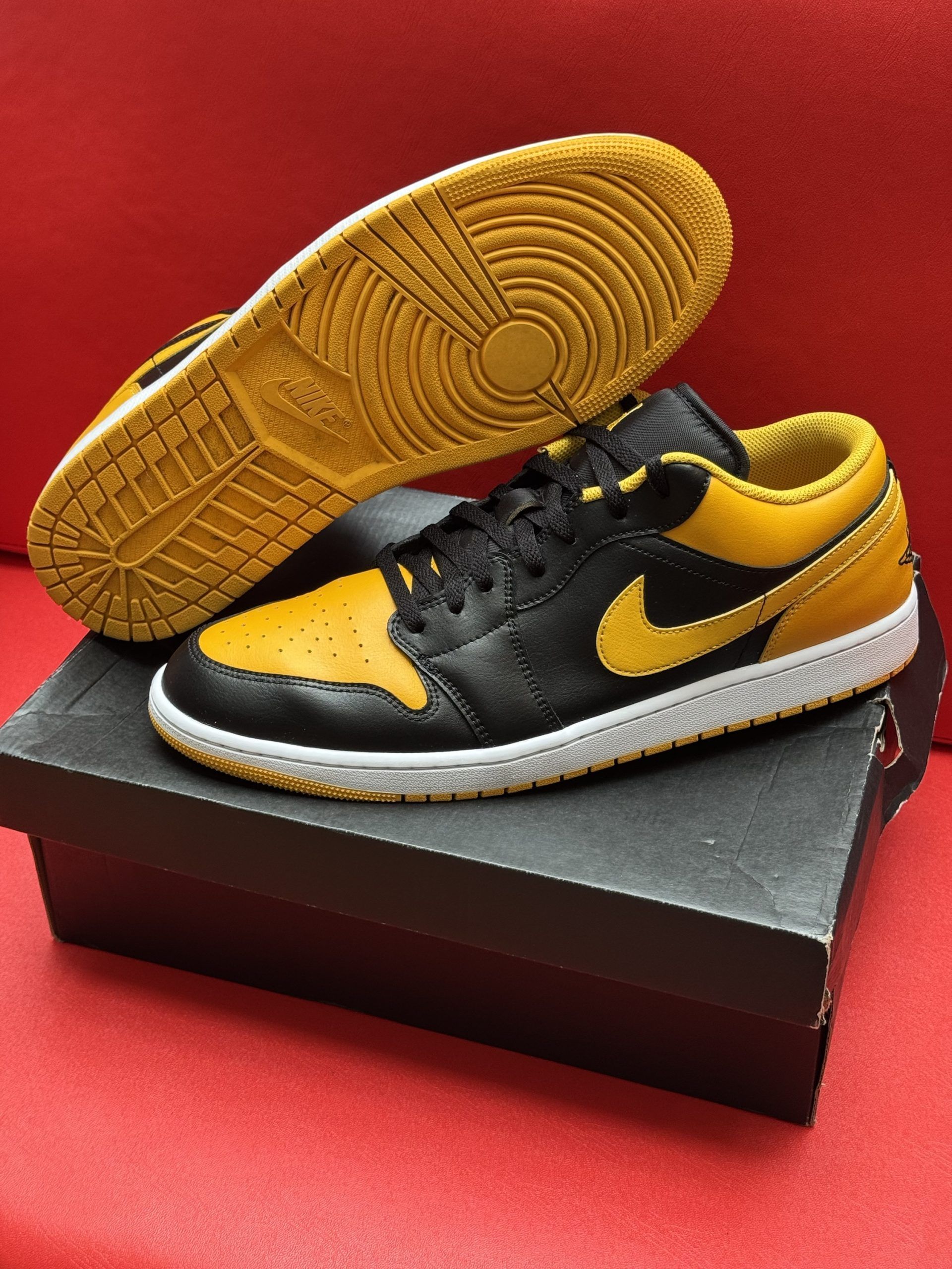 Black and yellow Air Jordan 1 Low sneakers with yellow soles, sitting on a black box, against a red backdrop.