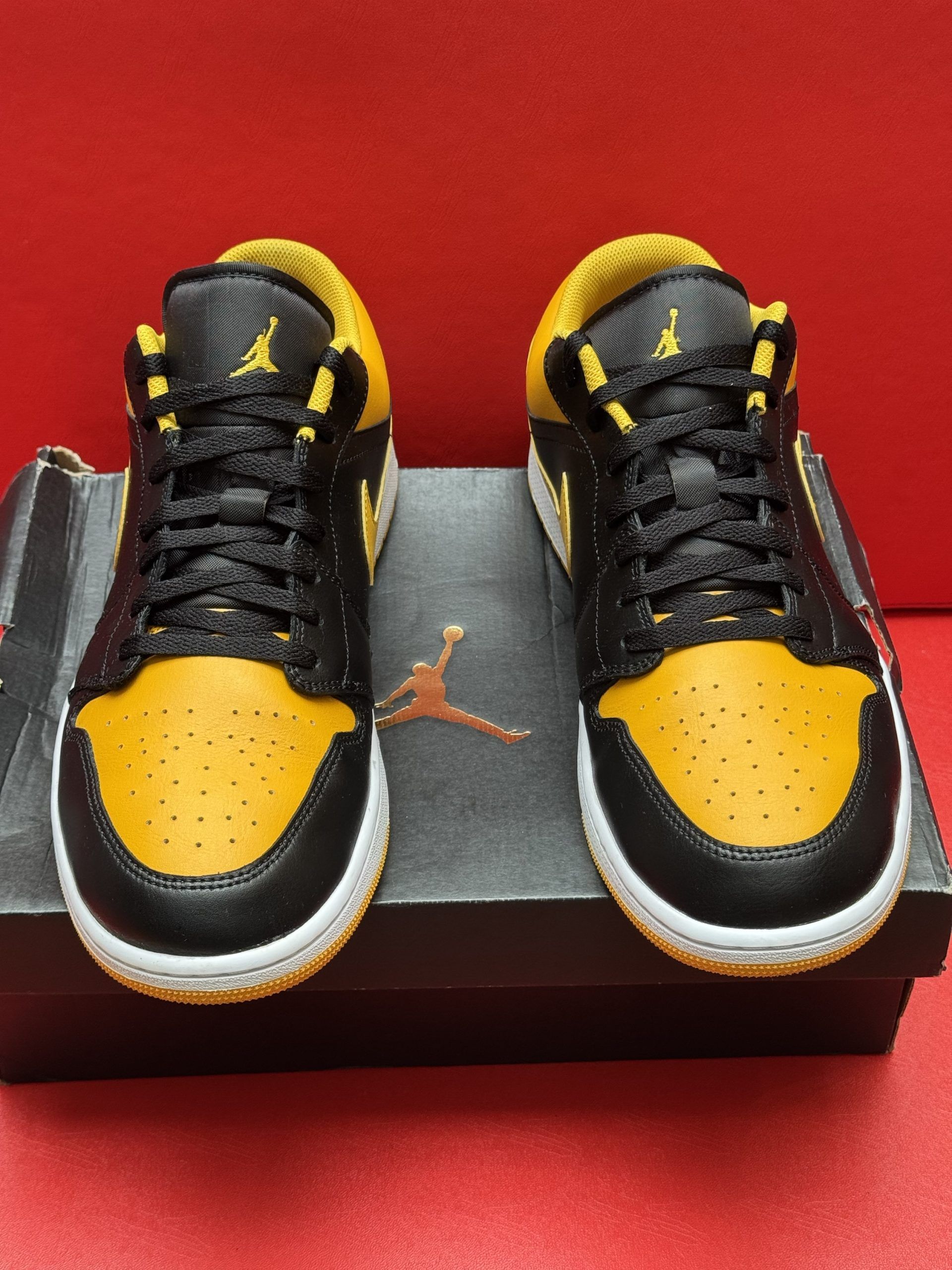 Black and yellow Air Jordan 1 Low sneakers with yellow soles, sitting on a black box, against a red backdrop.