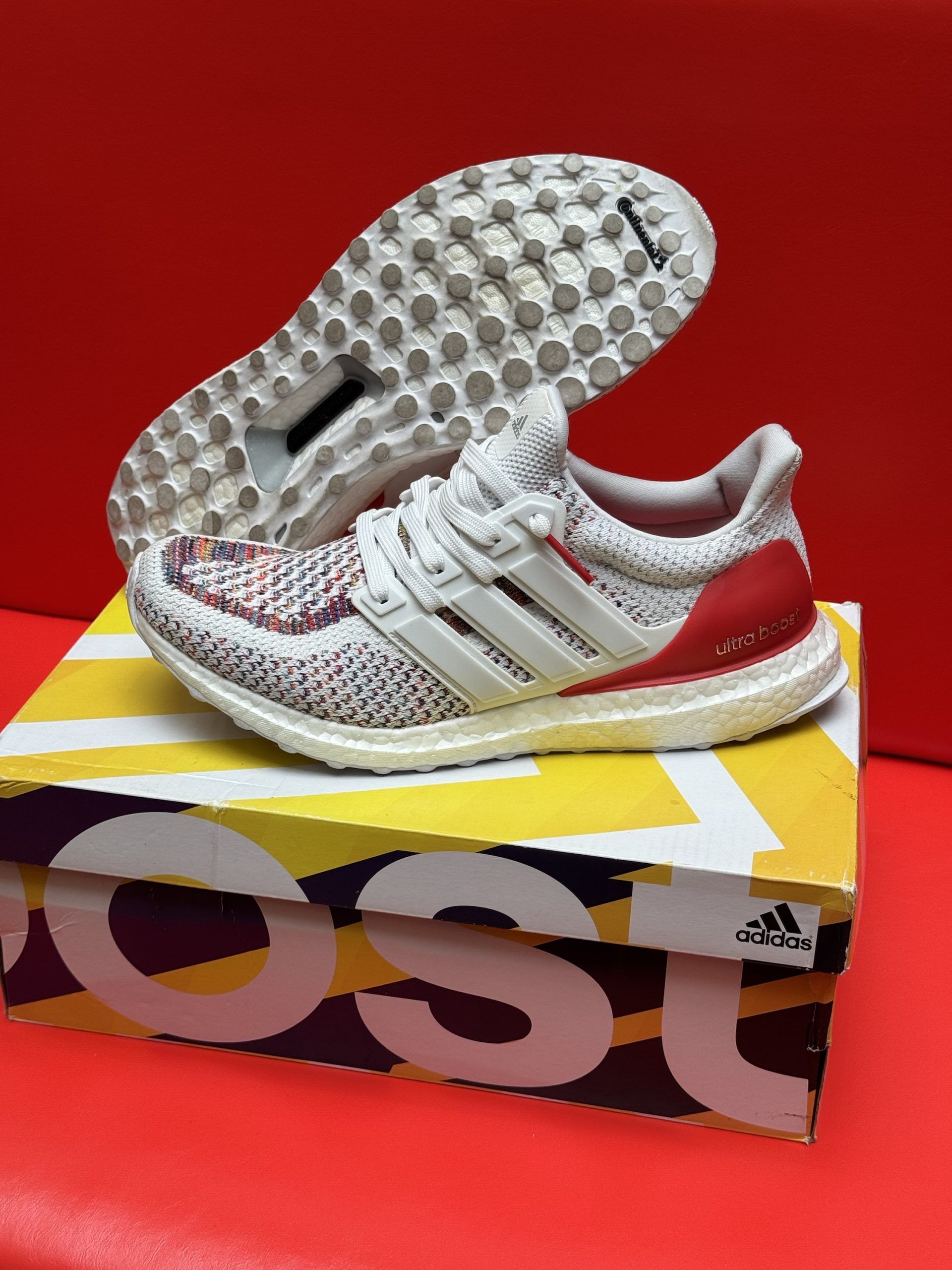 White and red Adidas Ultraboost sneakers on a box, with the sole visible, against a red background.