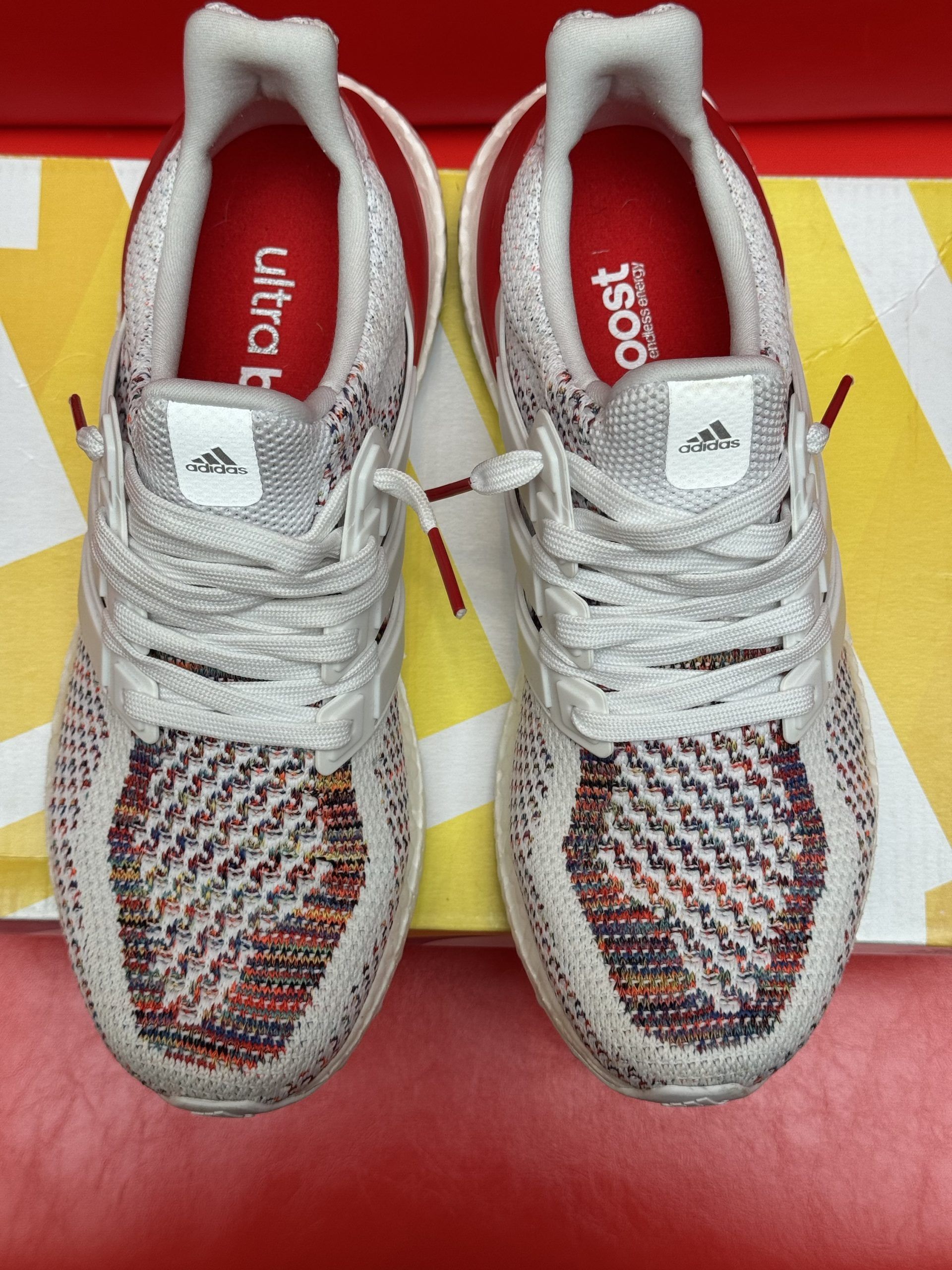White and red Adidas Ultraboost sneakers on a box, with the sole visible, against a red background.