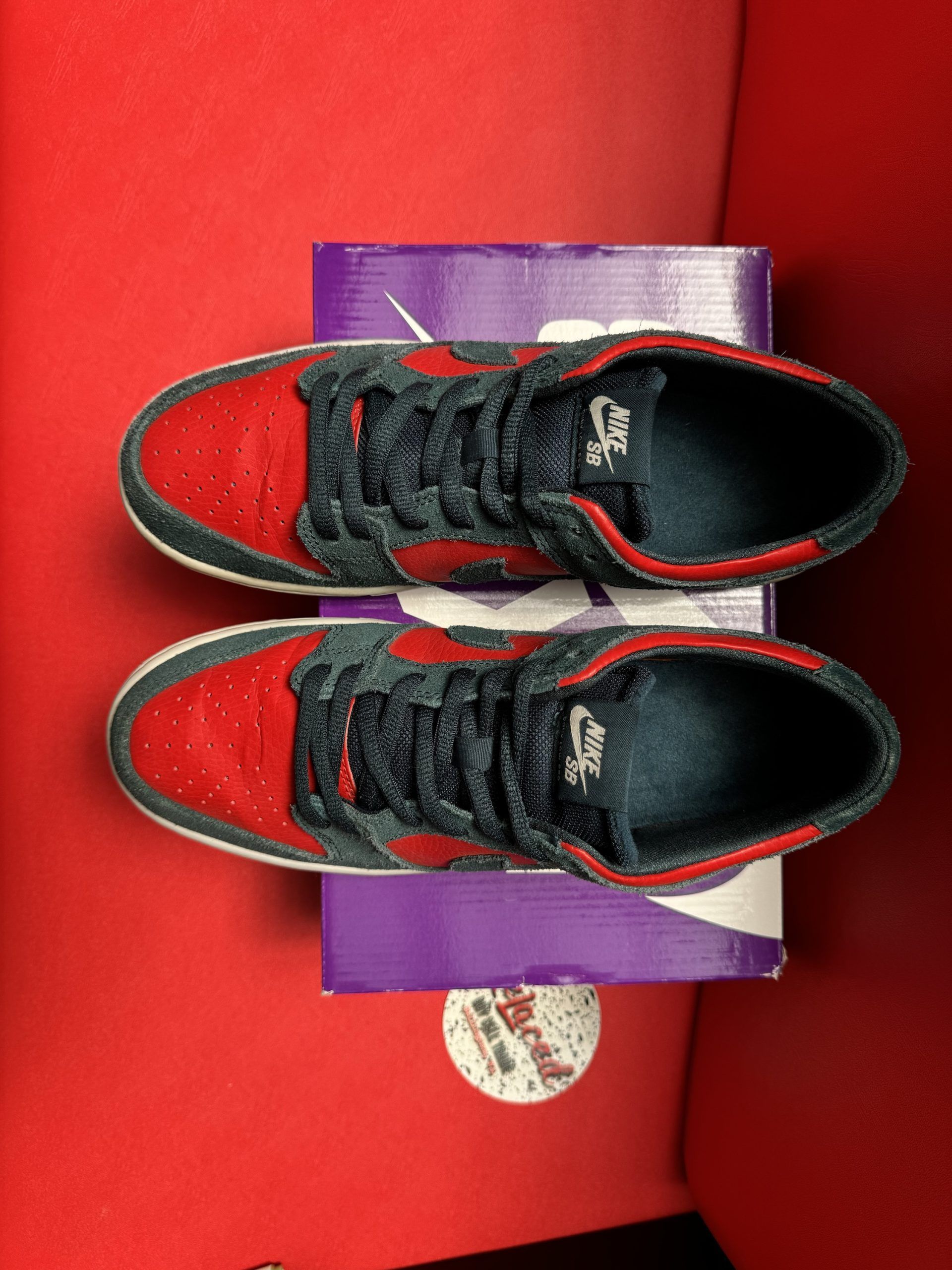 Red and black Nike Dunk SB sneakers on a purple box, one shoe upside down showing red sole.