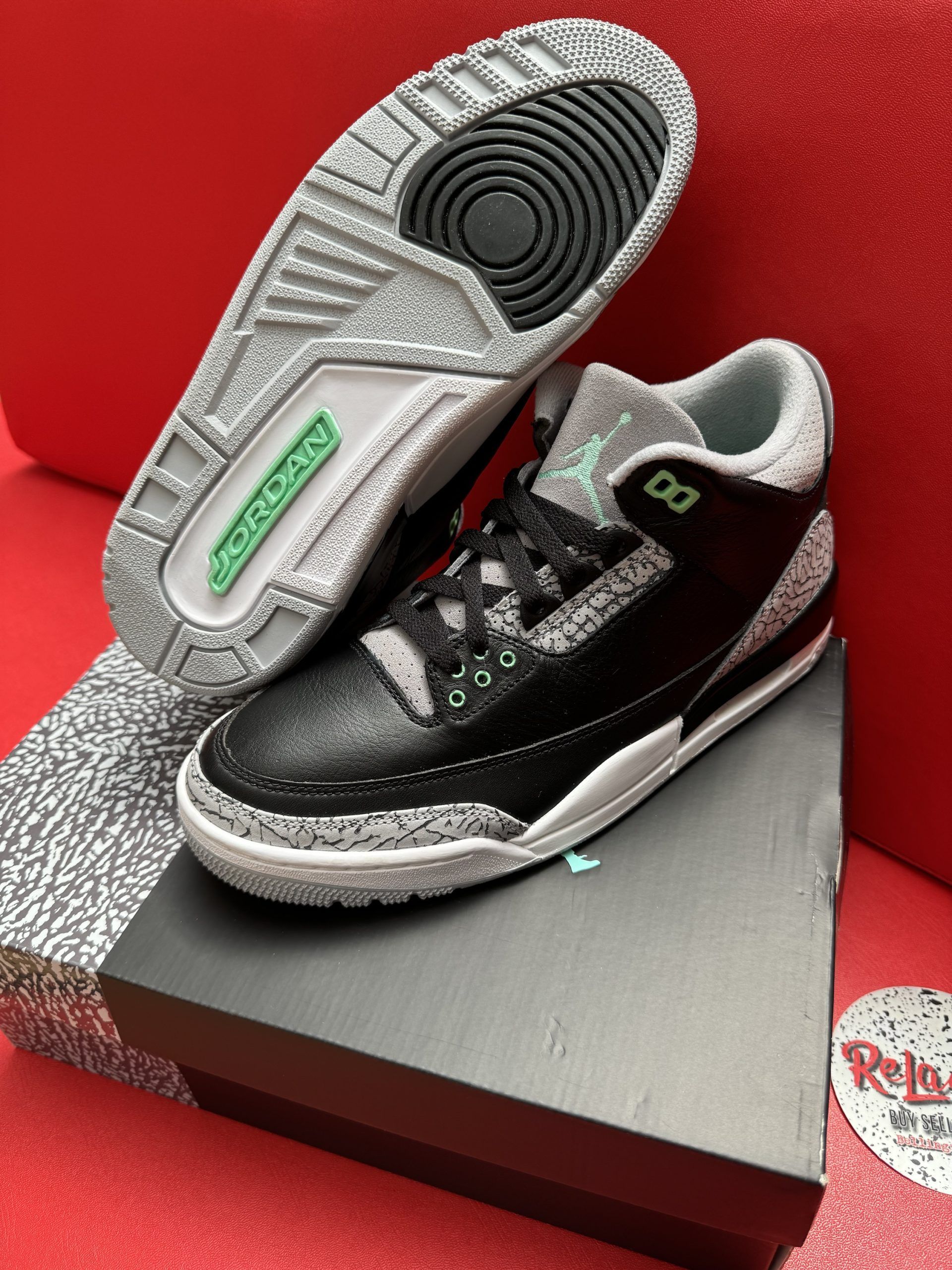 Black and gray Air Jordan 3 sneakers with green accents, displayed on a shoebox against a red backdrop.