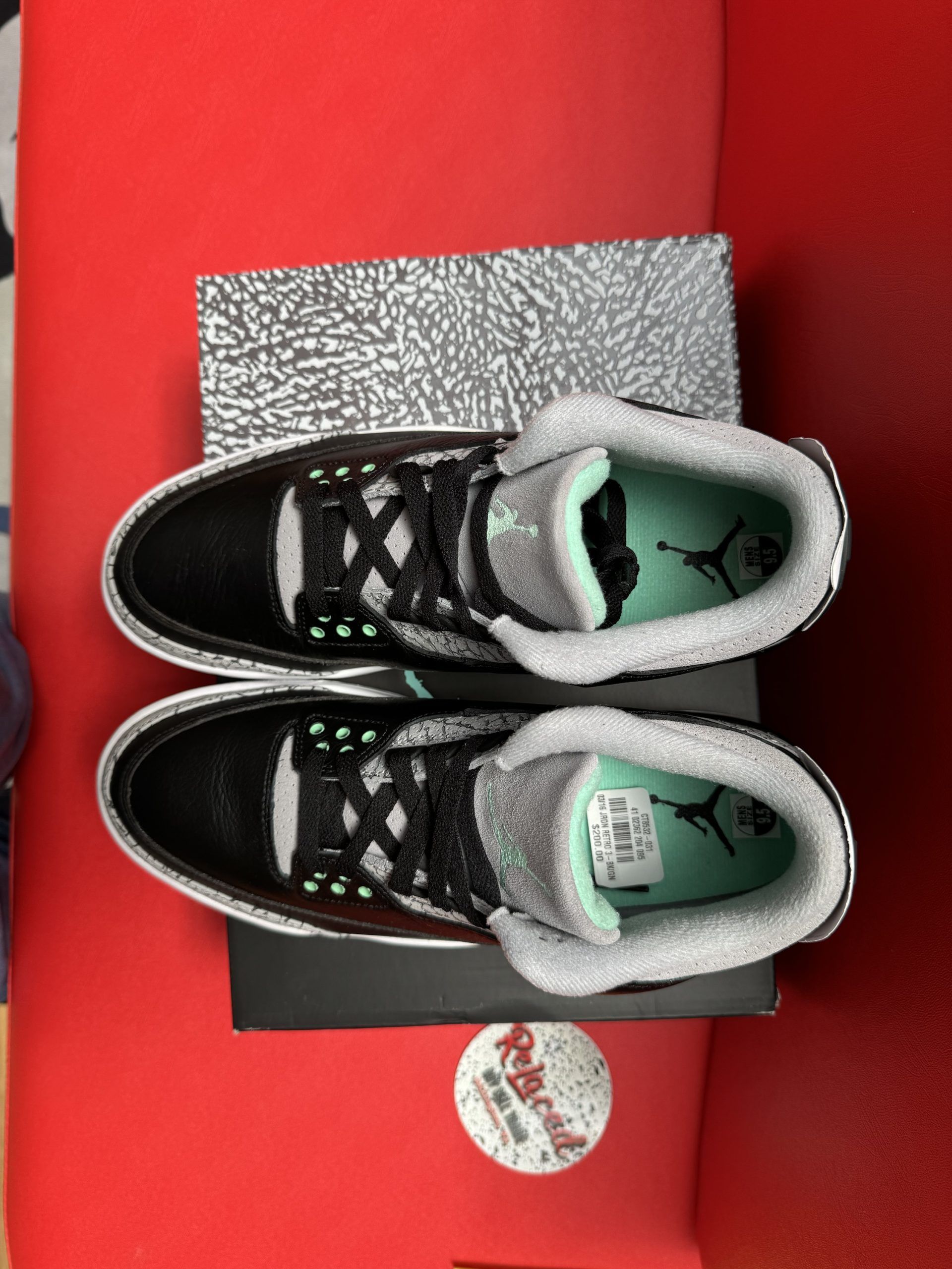 Black and gray Air Jordan 3 sneakers with green accents, displayed on a shoebox against a red backdrop.