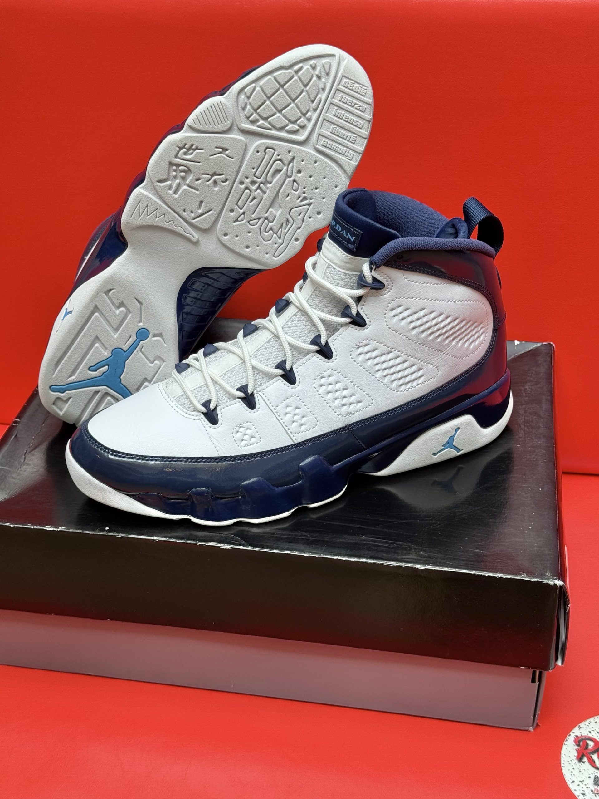 Air Jordan 9 sneakers in white and navy blue on a black box, set against a red background.