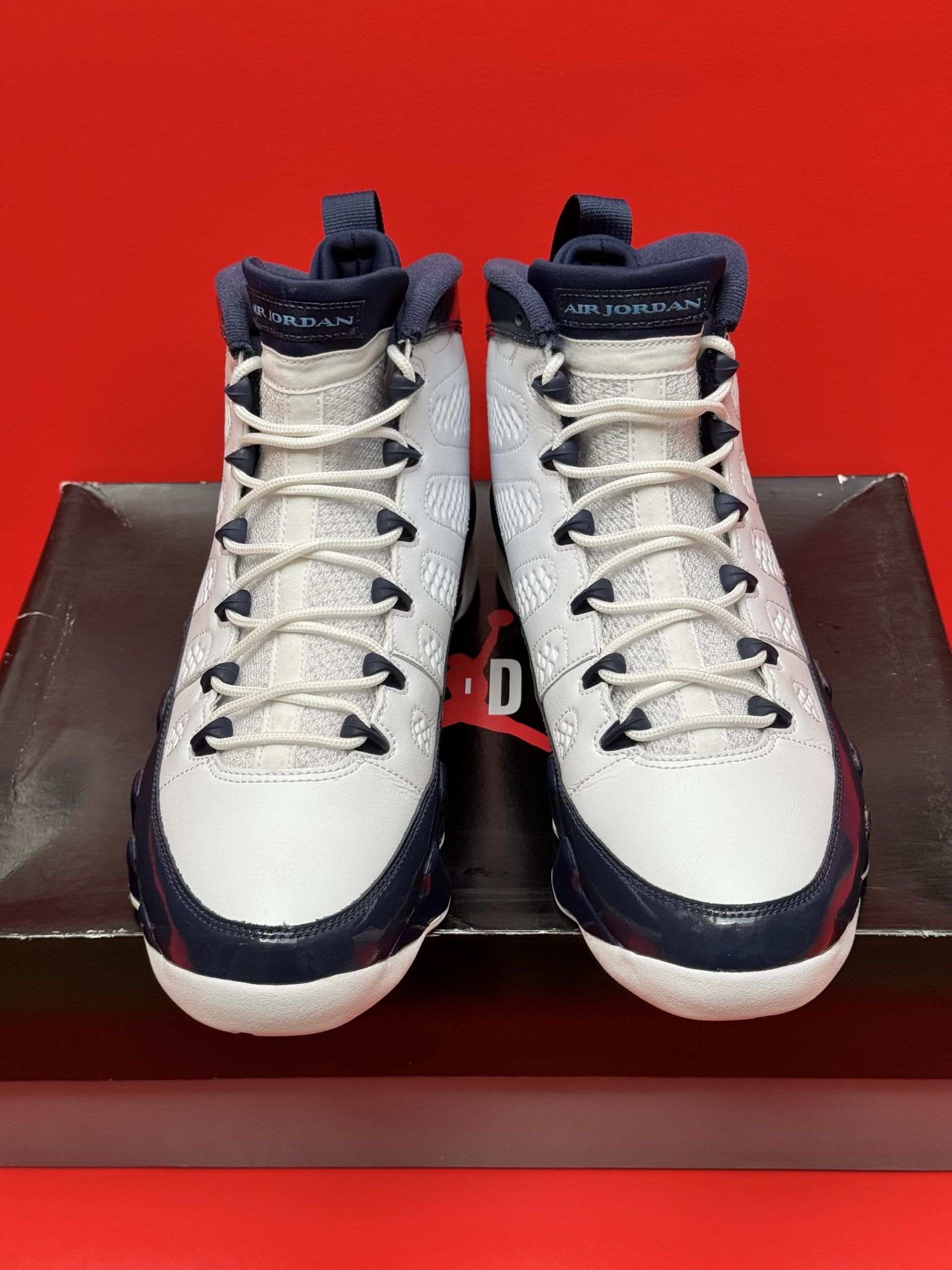Air Jordan 9 sneakers in white and navy blue on a black box, set against a red background.