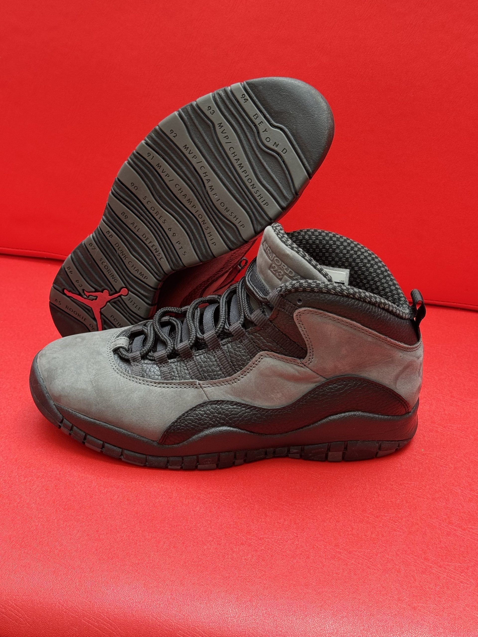 Gray and black Air Jordan 10 sneakers on a red surface. Sole is visible with red Jumpman logo.