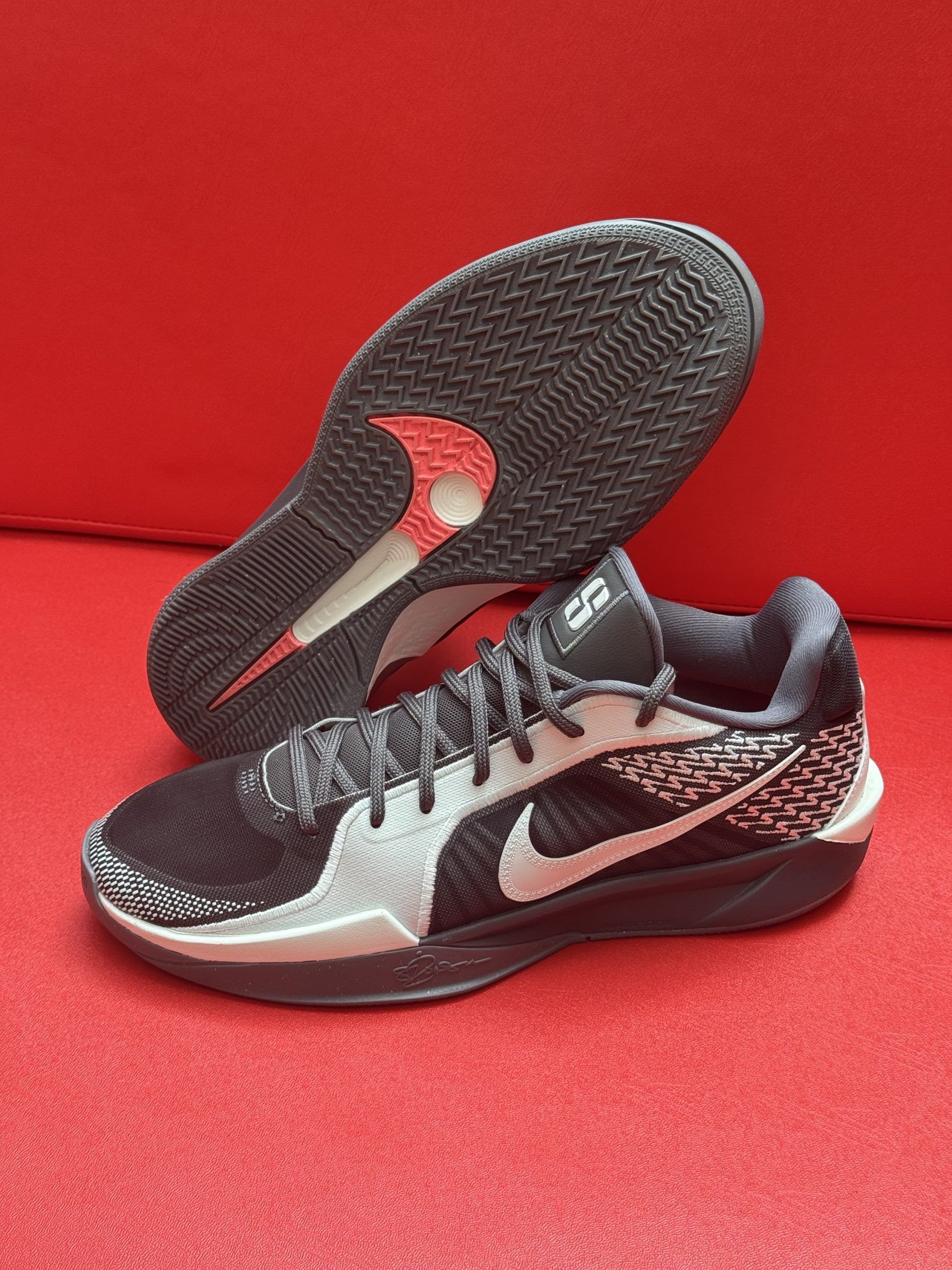 Black and white Nike sneakers on a red surface. The sole of one shoe is visible, showing a red accent.