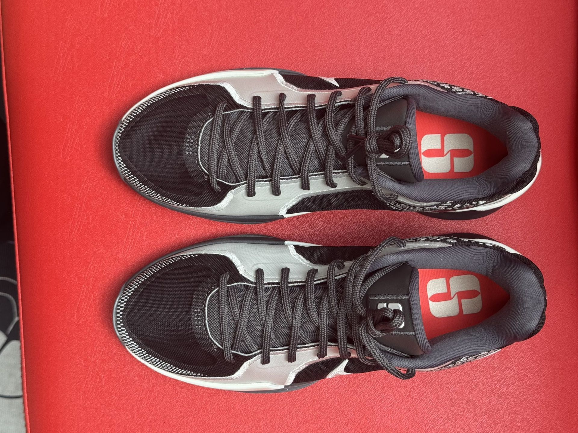 Black and white Nike sneakers on a red surface. The sole of one shoe is visible, showing a red accent.