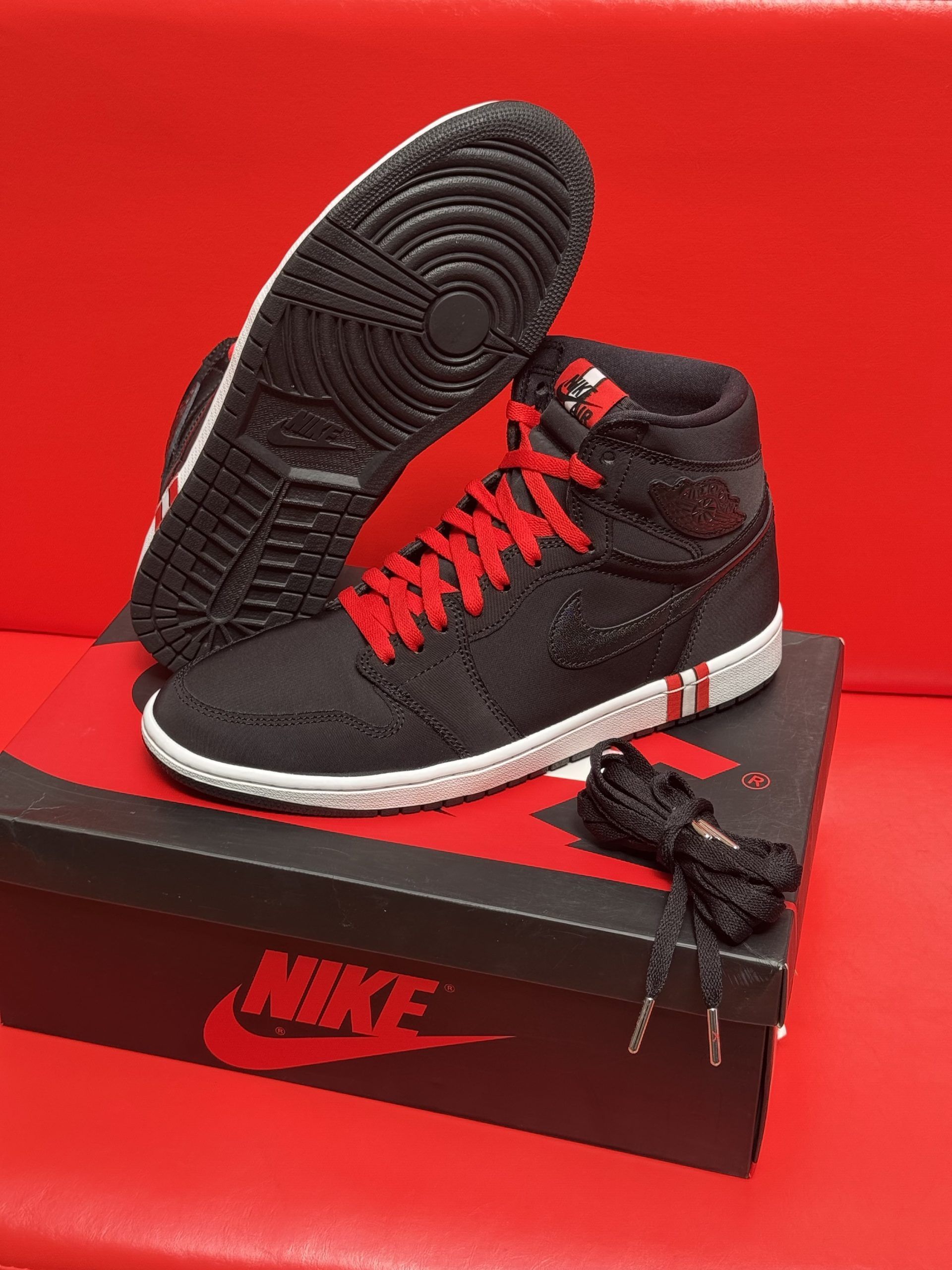 Black and red Air Jordan 1 high-top sneakers on a Nike box, with a red background. One shoe shows the sole.