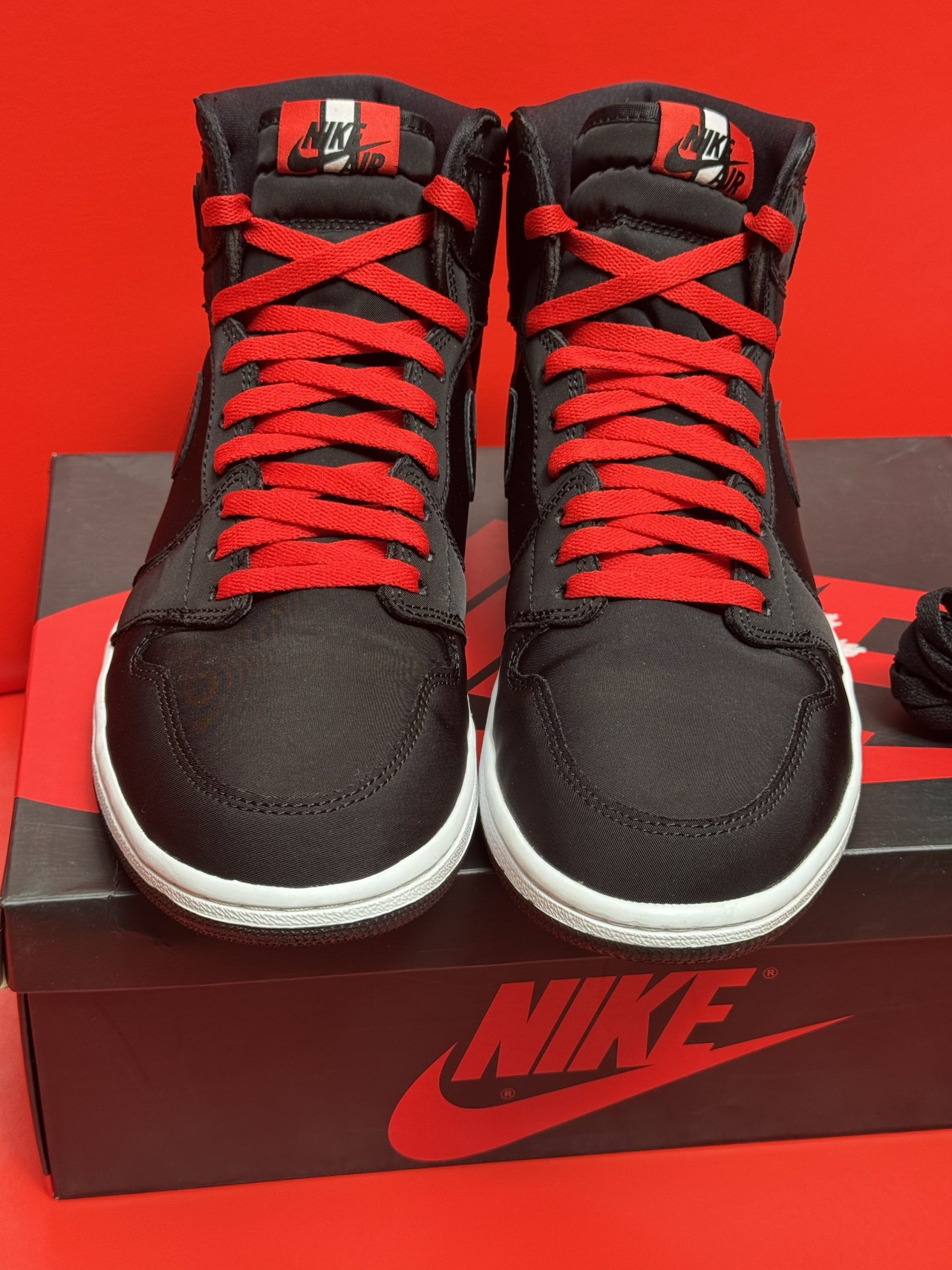 Black and red Air Jordan 1 high-top sneakers on a Nike box, with a red background. One shoe shows the sole.