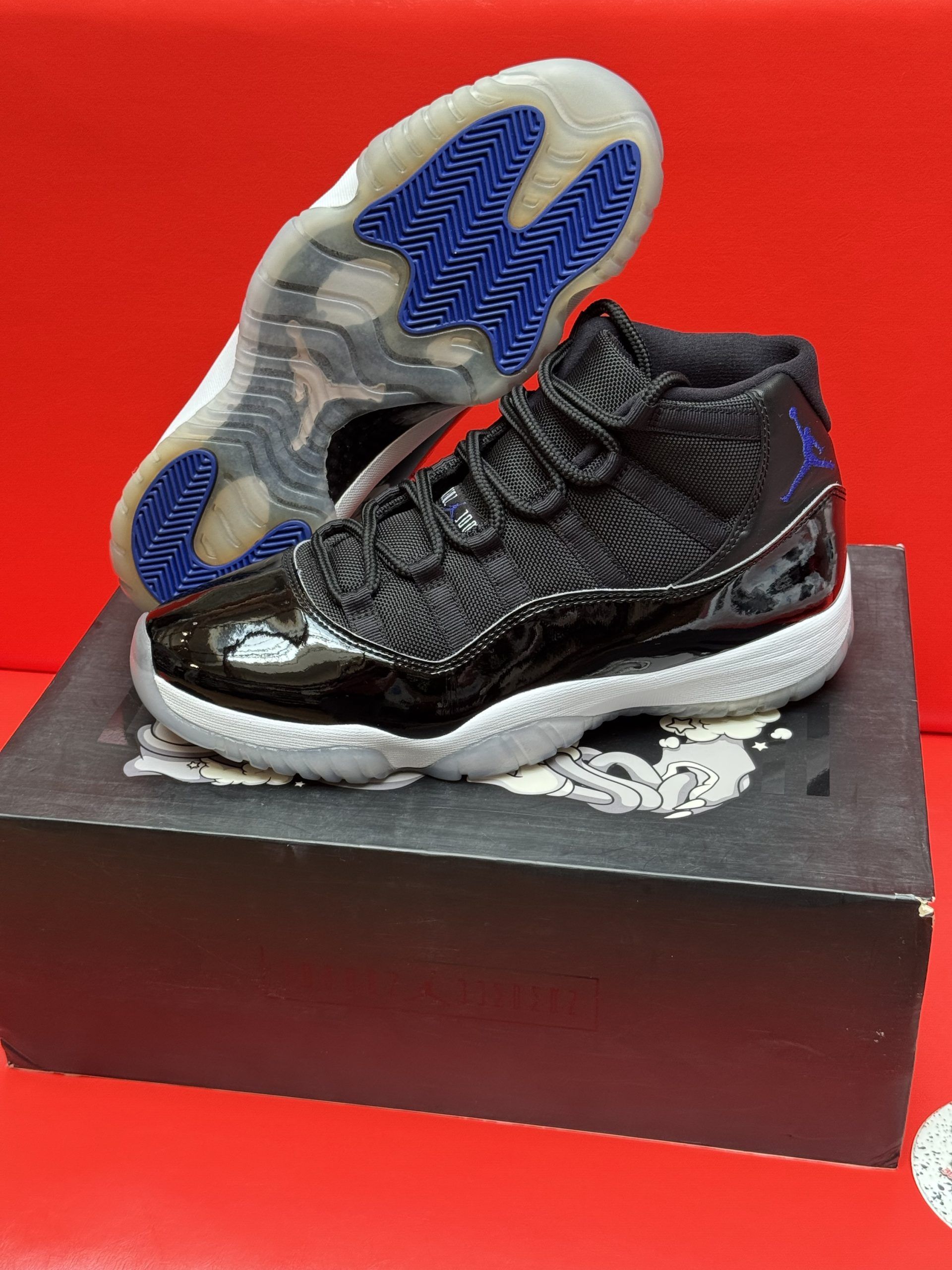 Black and white Air Jordan 11 sneaker on a box, with a blue and white sole against a red backdrop.