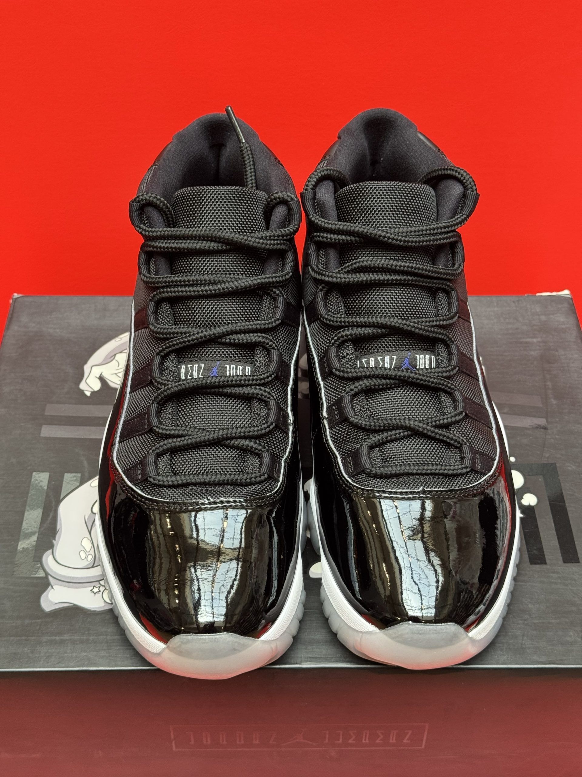 Black and white Air Jordan 11 sneaker on a box, with a blue and white sole against a red backdrop.