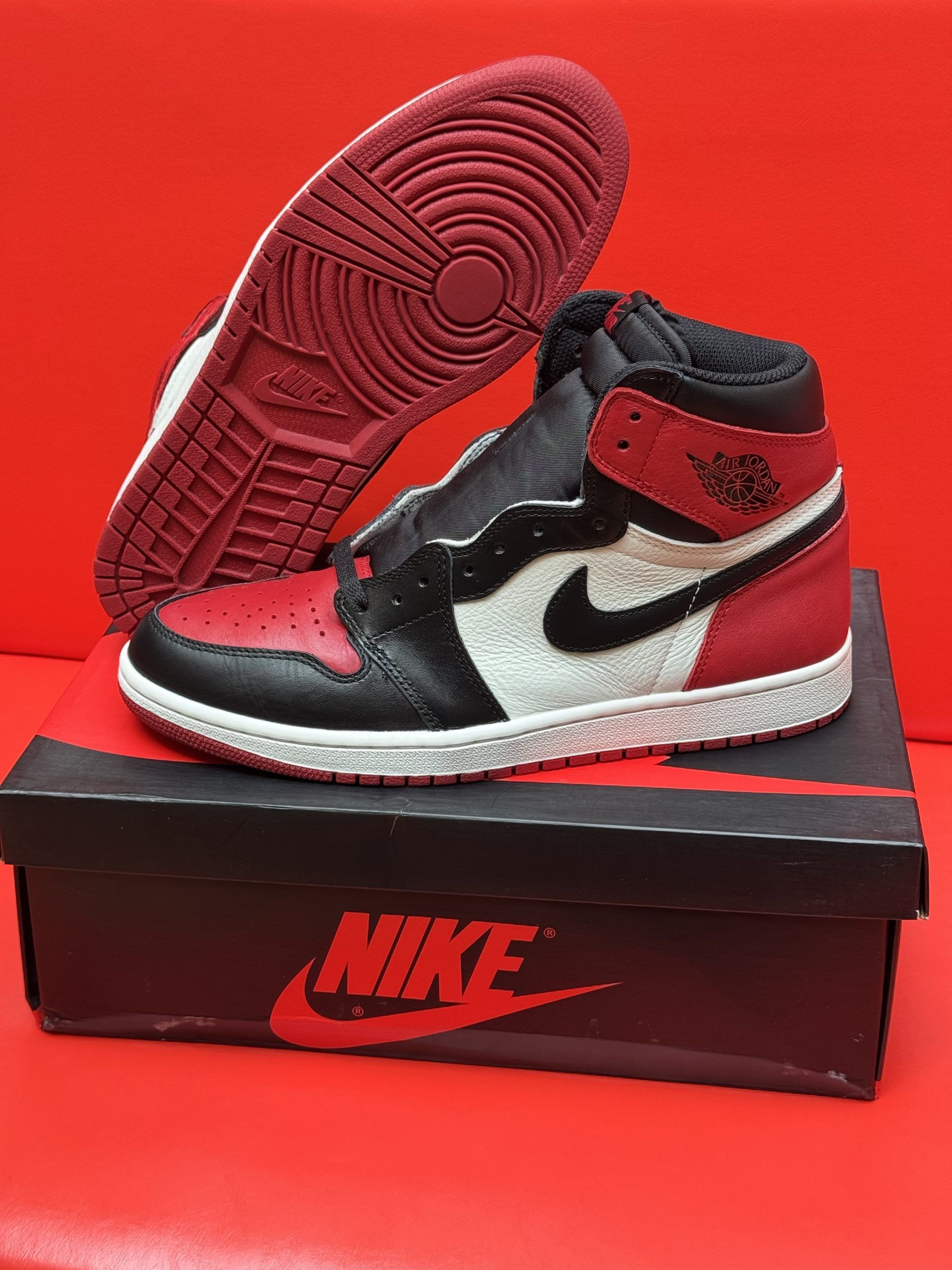 Air Jordan 1 sneakers in black, white, and red on a shoebox against a red background.
