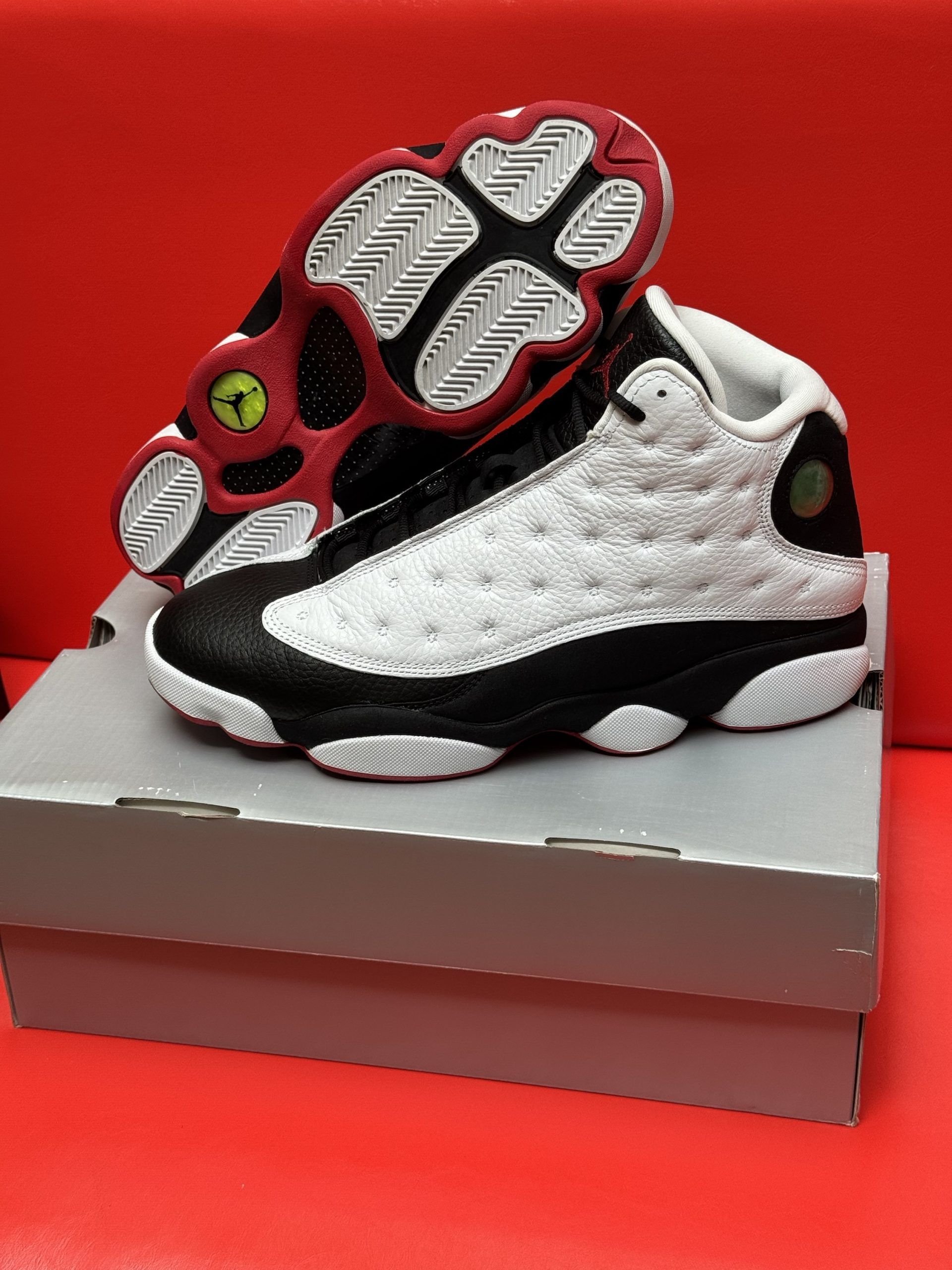 Pair of white, black, and red Jordan 13 sneakers on a grey shoe box, set against a red background.